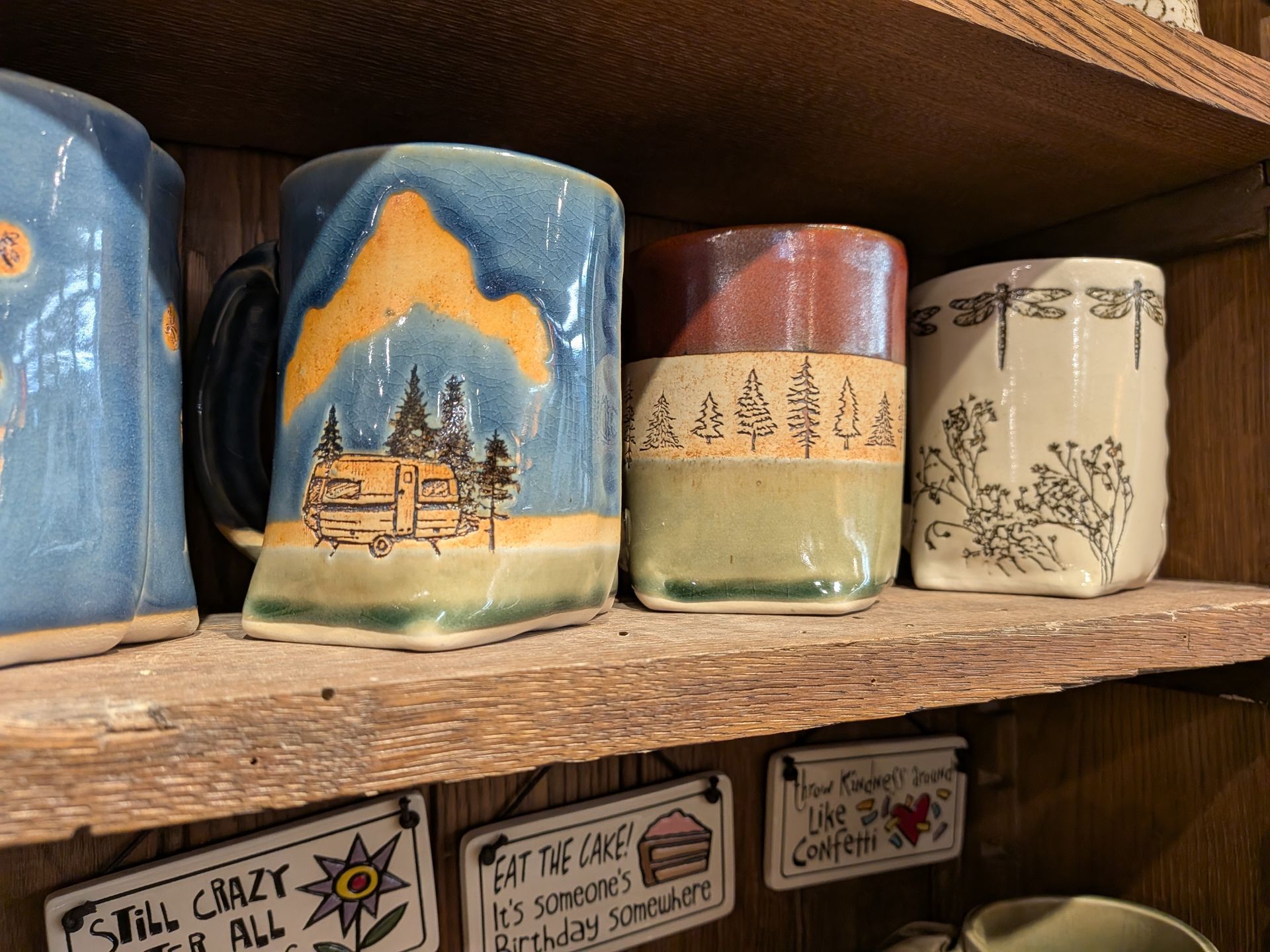Painted ceramic mugs on a wooden shelf, including a blue mountain scene mug and floral designs.