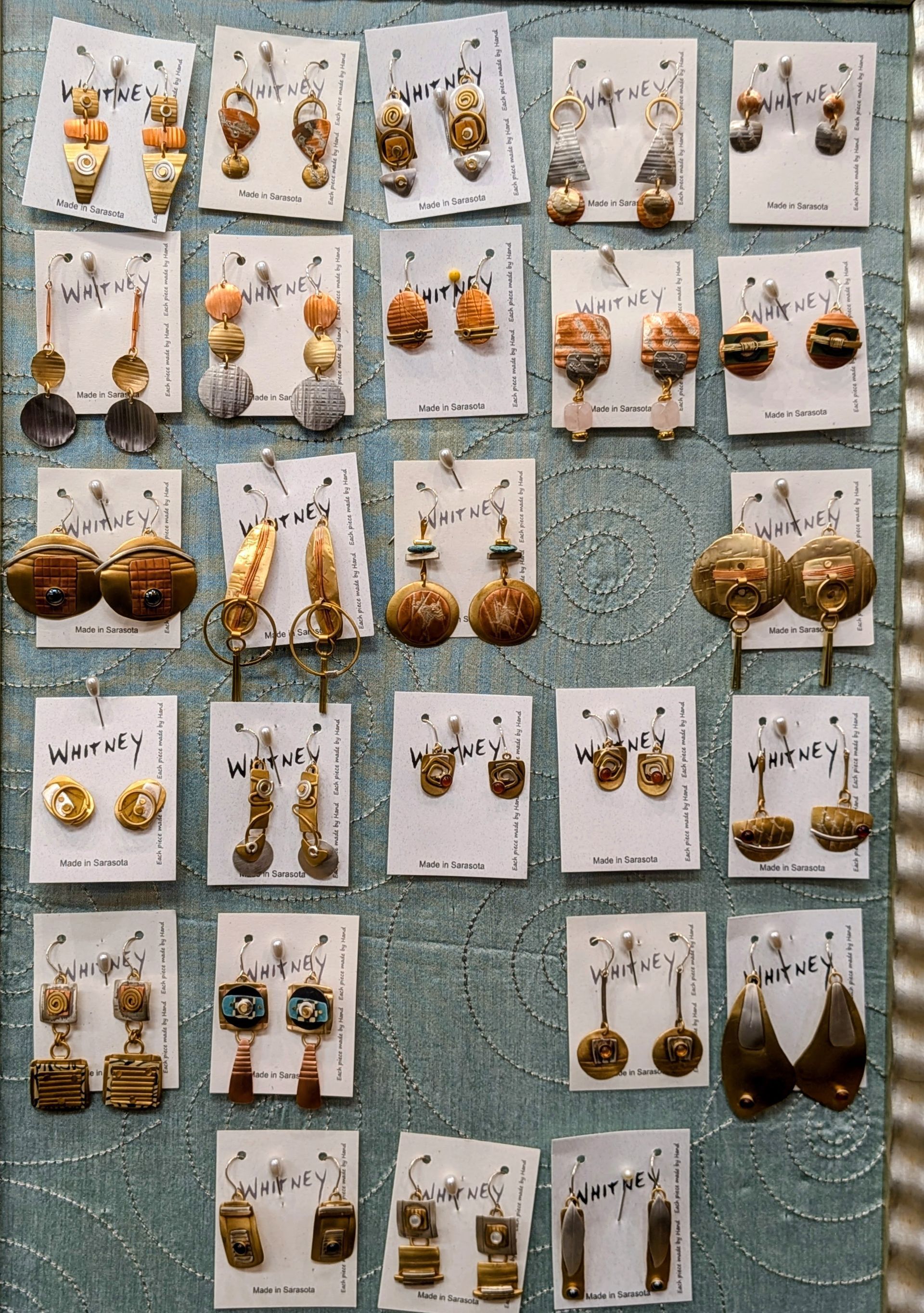 Display of handmade earrings on white cards pinned to a blue textured board.