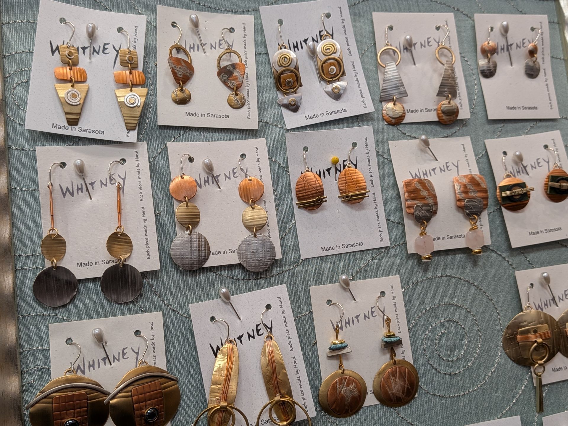 Assorted handmade earrings displayed on white cards against a teal patterned surface.