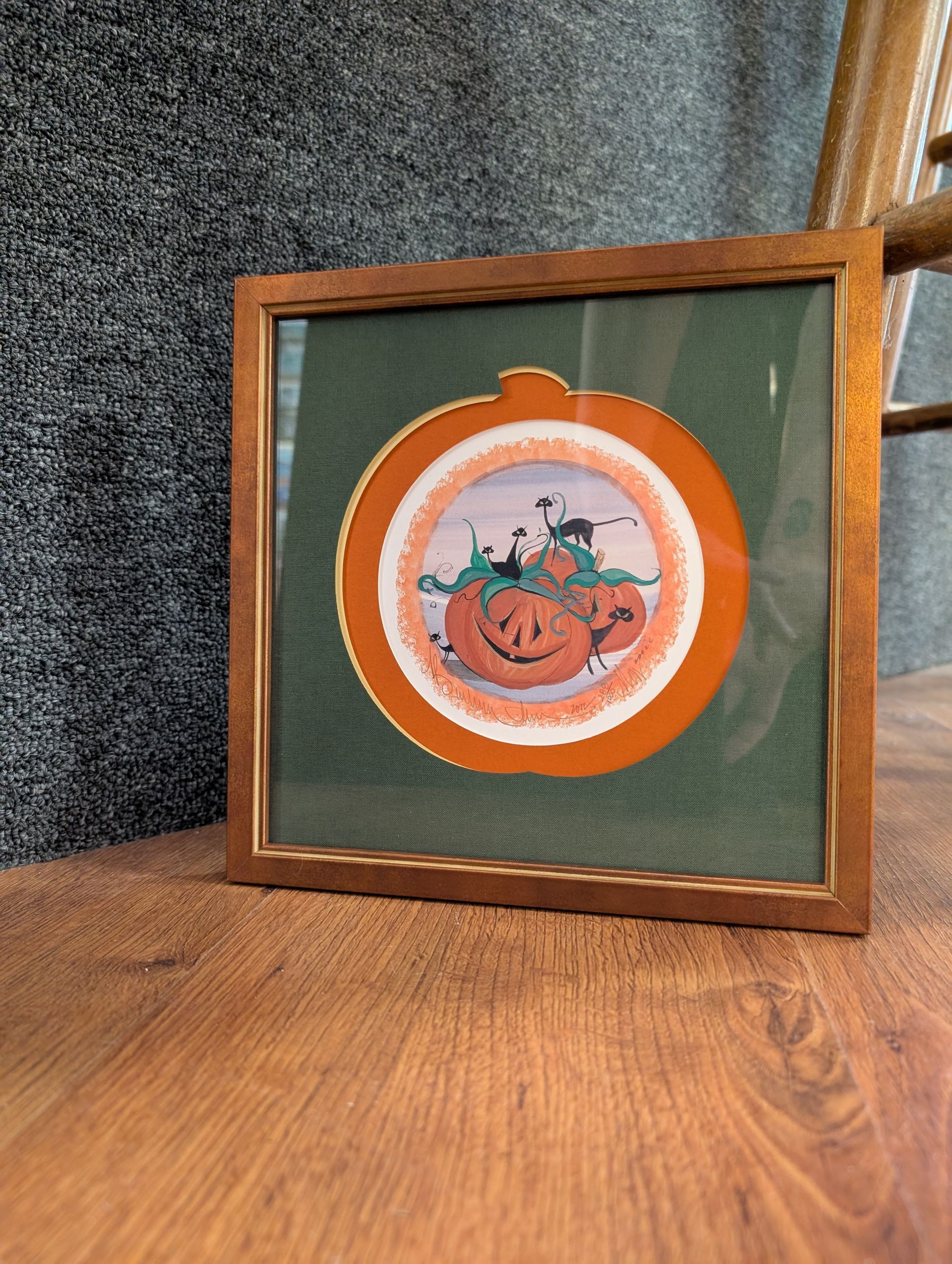 Framed autumn pumpkin illustration on a green background, resting on a wooden surface.
