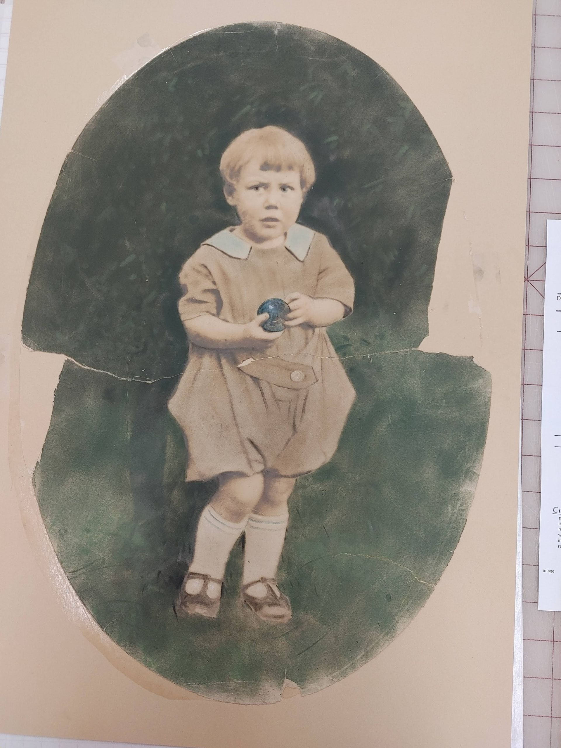 Child in a tan outfit holding a small object, standing against a dark green oval background.