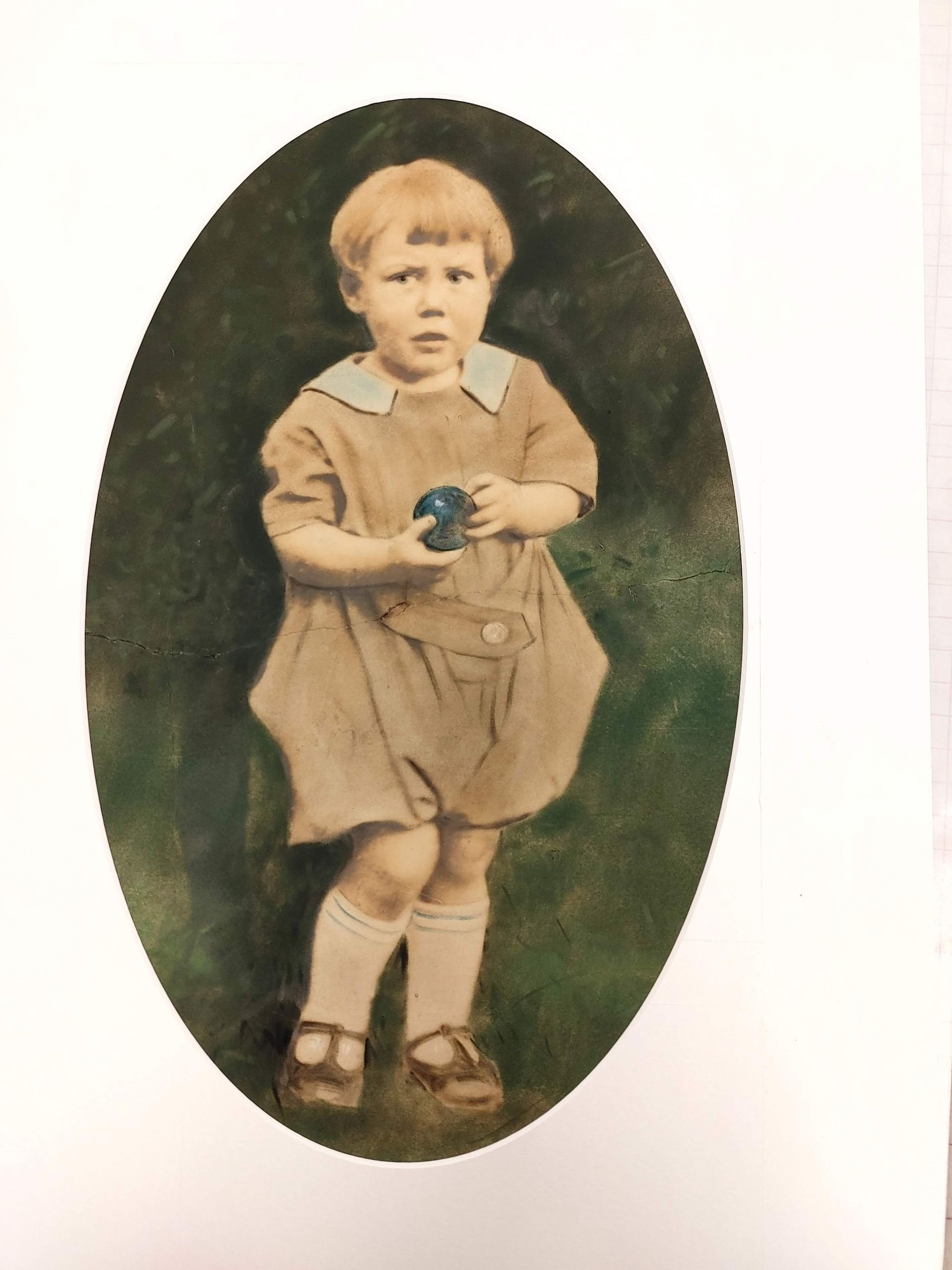 Sepia-toned portrait of a child standing in an oval frame, holding a small object, wearing a dress and boots.