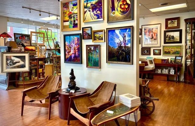 Interior of an art gallery featuring multiple paintings hung on white walls, two wooden chairs, and a small table.