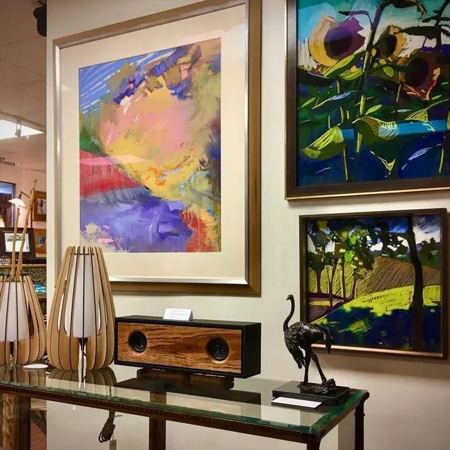 Art gallery interior with three framed paintings, a wooden speaker, two lamps, and a small ostrich sculpture on a table.