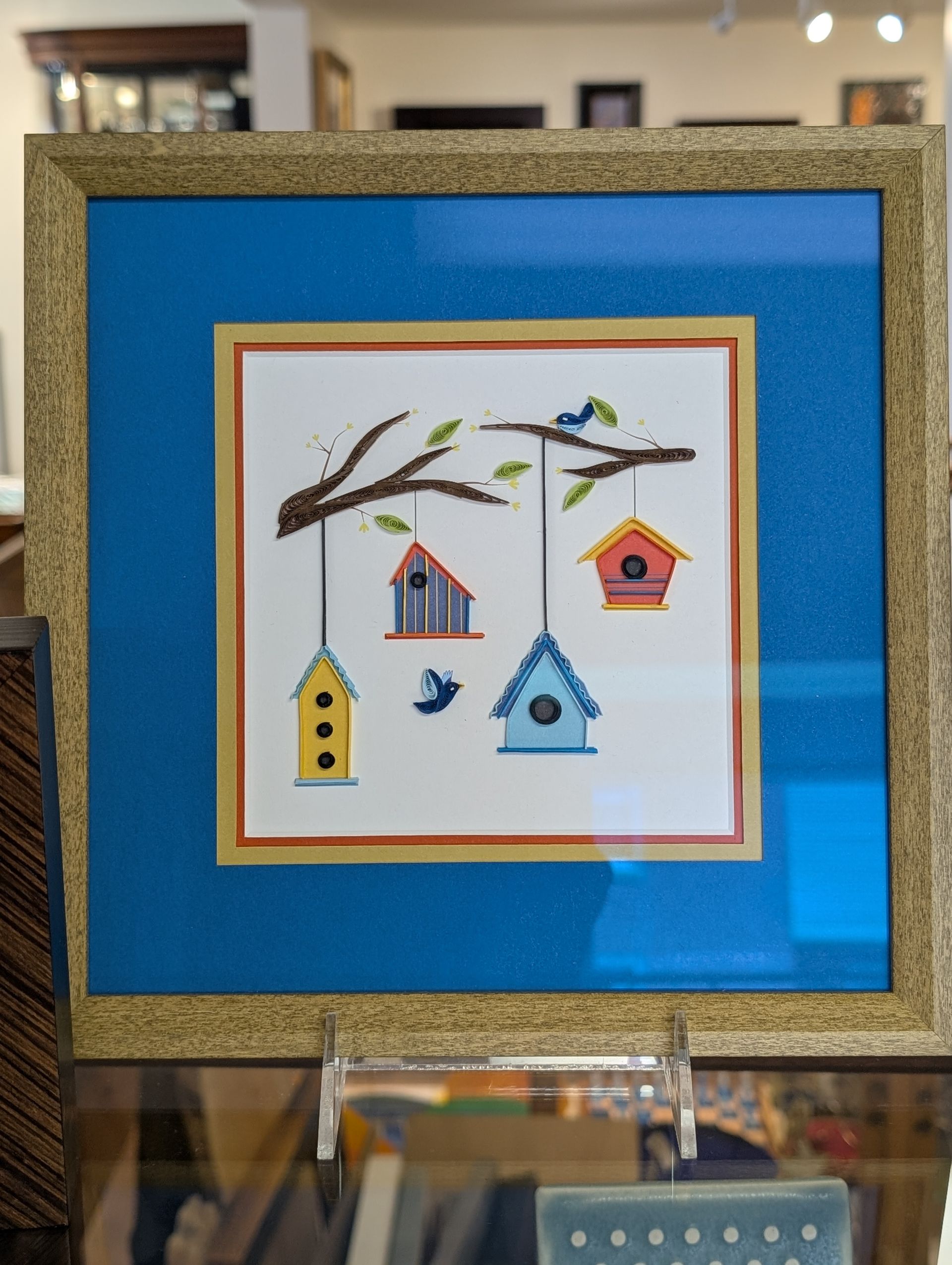 Framed birdhouse artwork on a blue mat, showing colorful birdhouses and birds on a branch.