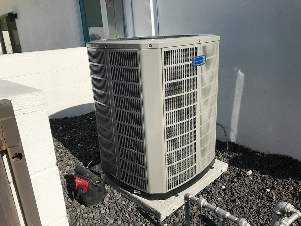 Air conditioner unit on a concrete base in an outdoor setting. Black rocks and a toolbox are nearby.