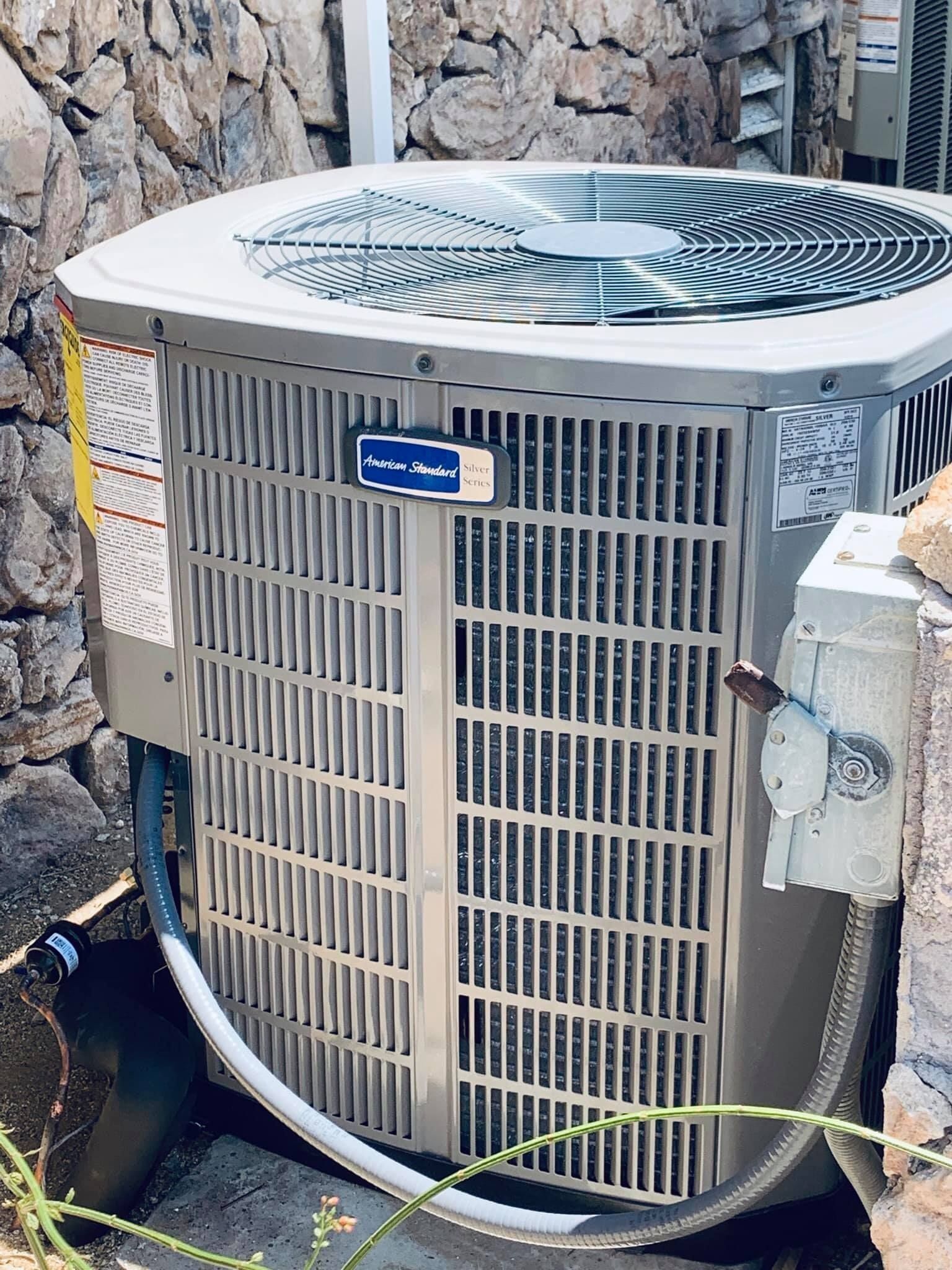 An American Standard AC unit next to a stone wall; beige, with a metal grate and fan visible.