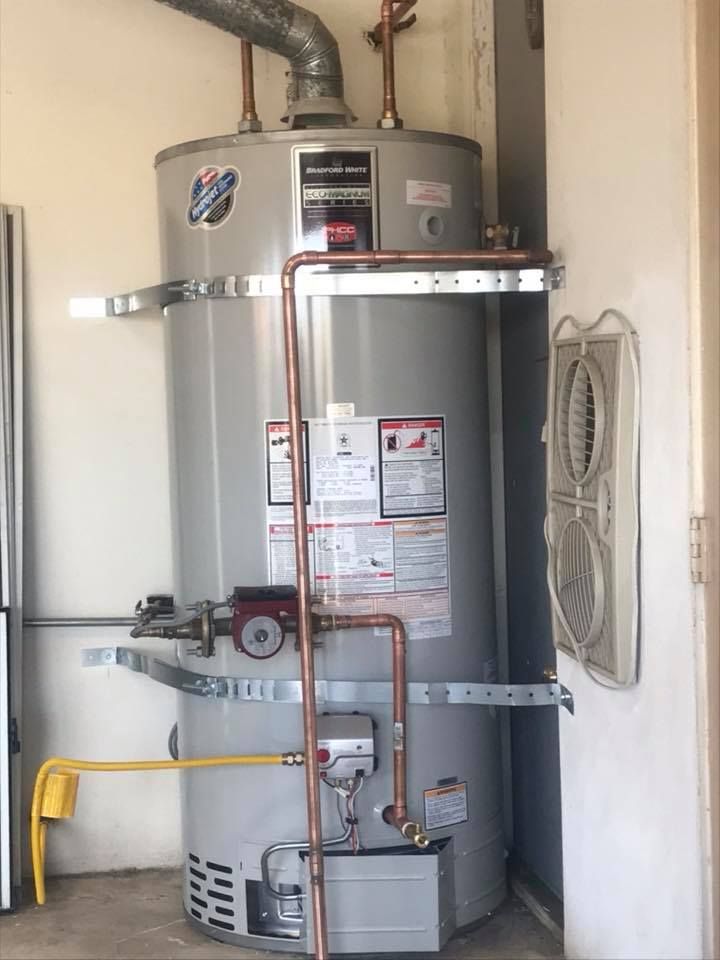 A tall, gray water heater with copper pipes and brackets is mounted on a garage wall next to a fan.
