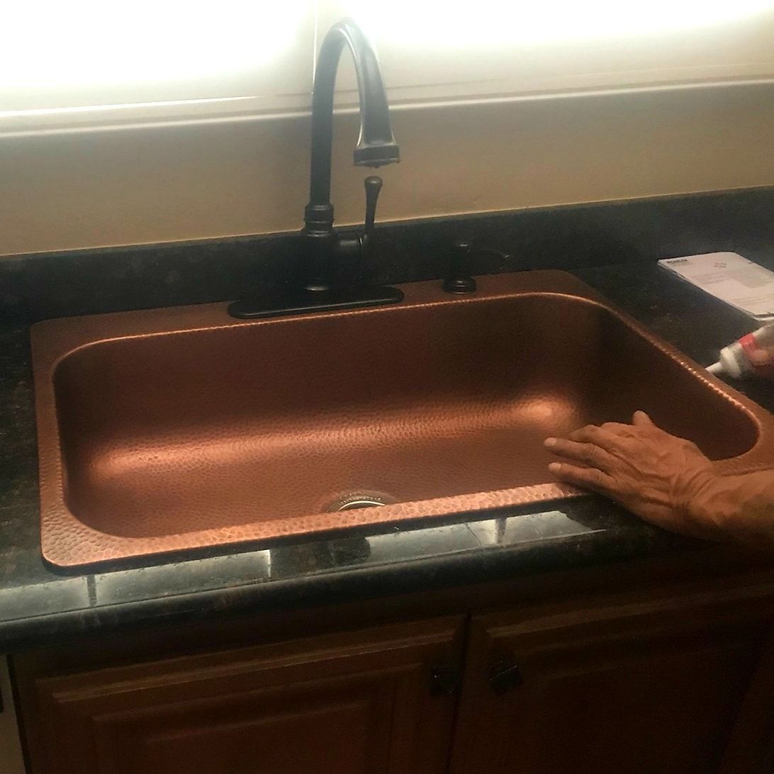 Copper kitchen sink with black faucet and countertop, person’s hand visible on the right side.