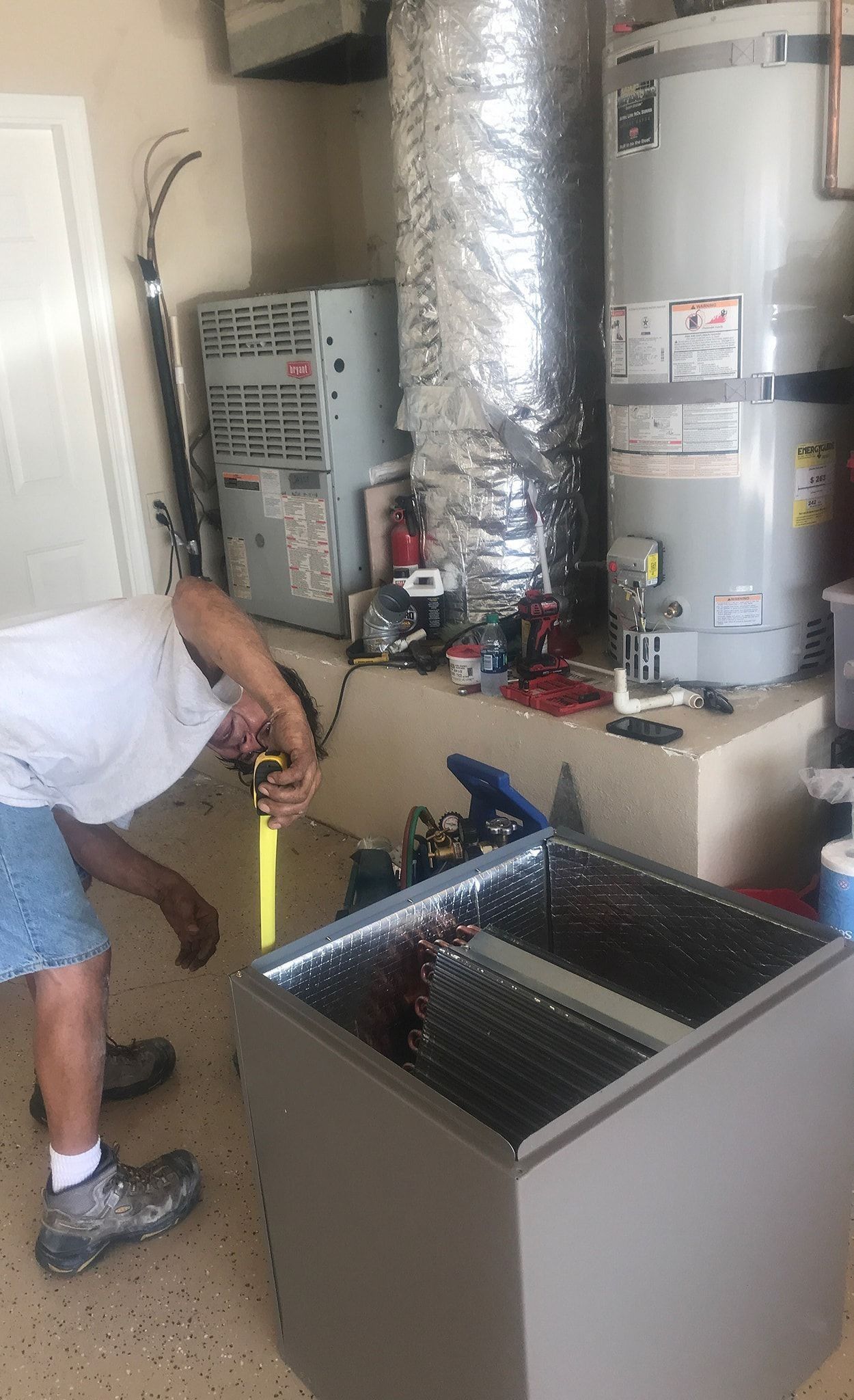 Man inspecting an air conditioning unit in a garage. He's wearing shorts and a white shirt.