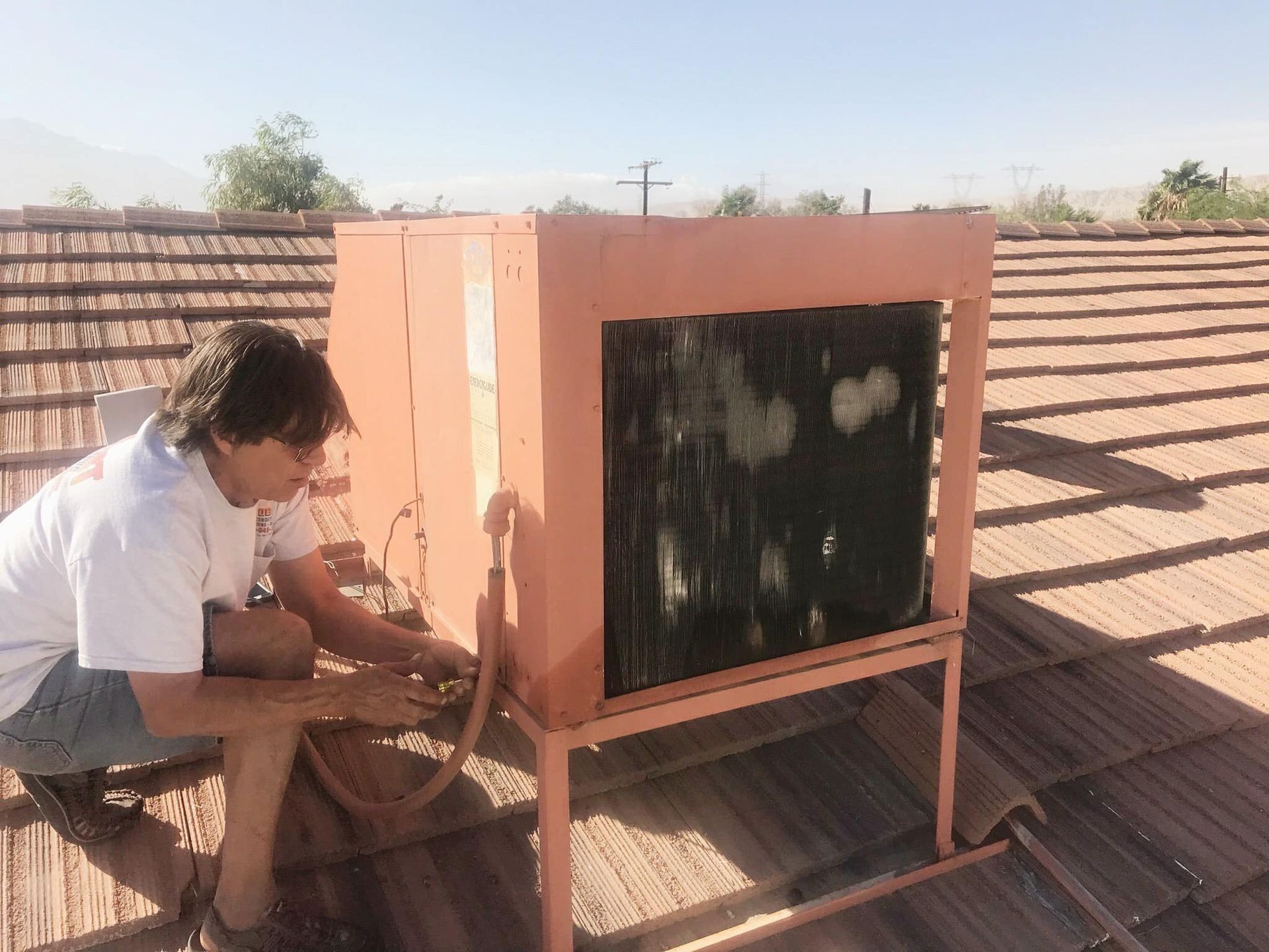 Person repairs an evaporative cooler on a rooftop; the cooler is peach-colored and against a mountain backdrop.