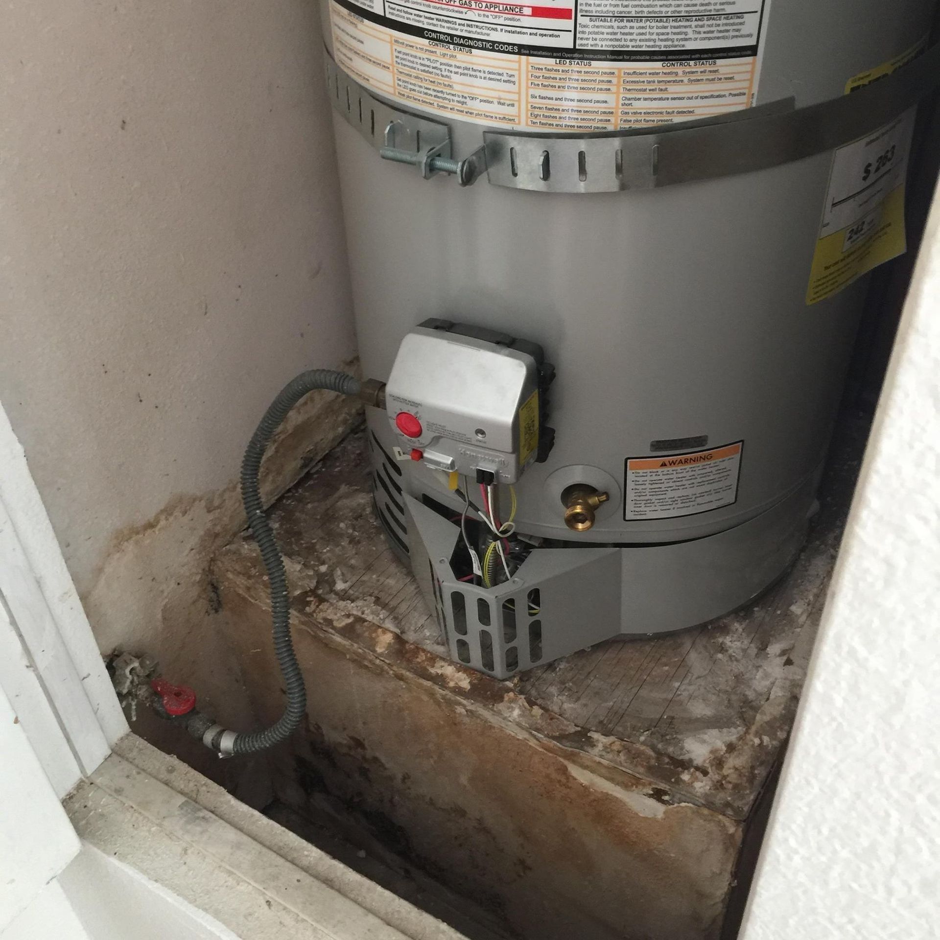 Water heater in a small enclosure; gray tank, gas line, control panel, white walls, concrete base.