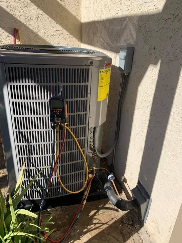 HVAC unit with gauges attached, outdoors. Gray, with yellow and black tubing. Located by a beige wall.