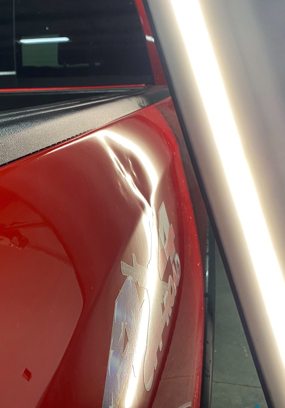 Red truck bed with a dent and peeling paint, illuminated by a light panel - before.