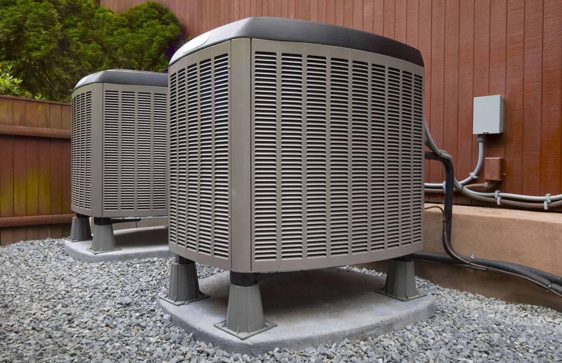 Two air conditioners are sitting on gravel outside of a house.