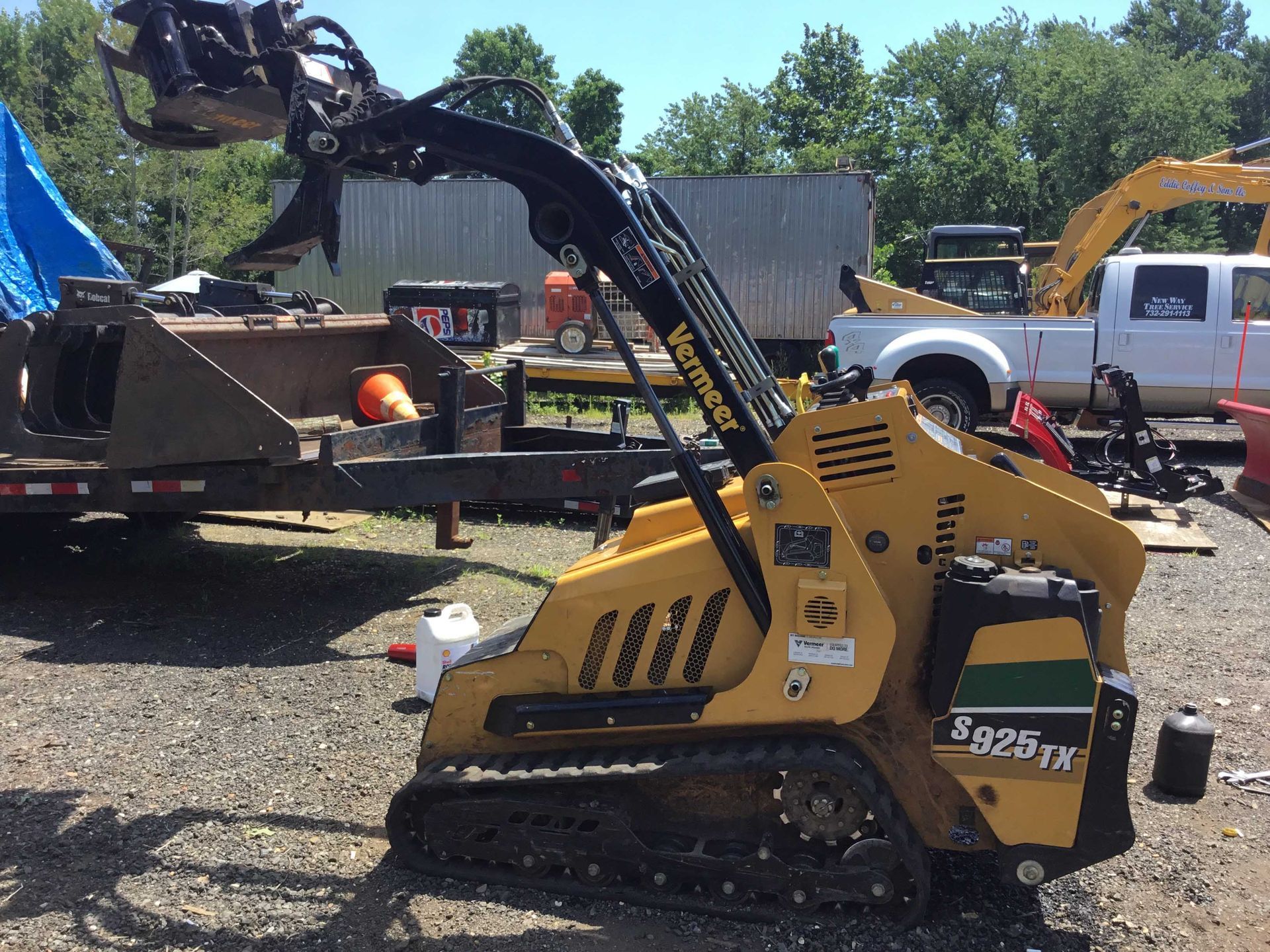 Yellow compact track loader with a black arm lifting debris in a sunny outdoor setting.