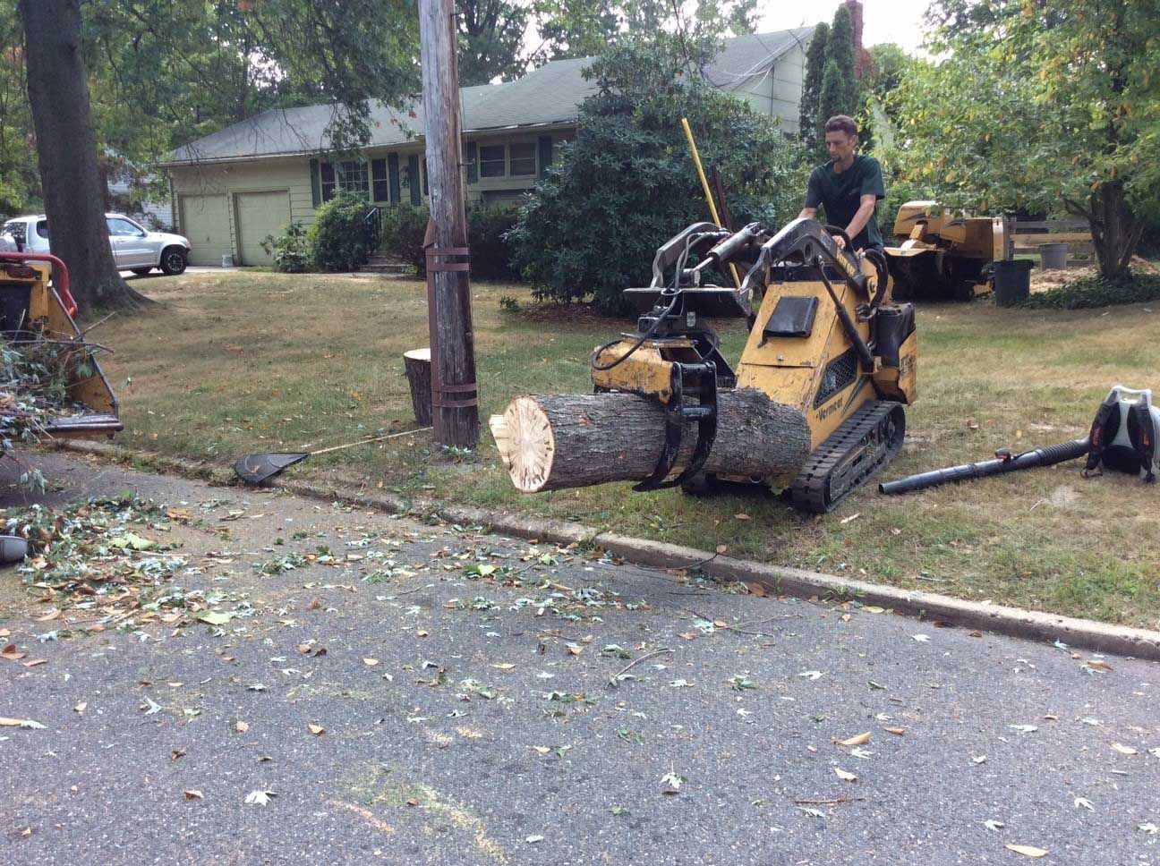 Man operating a yellow skid steer lifting a large tree trunk on a street.
