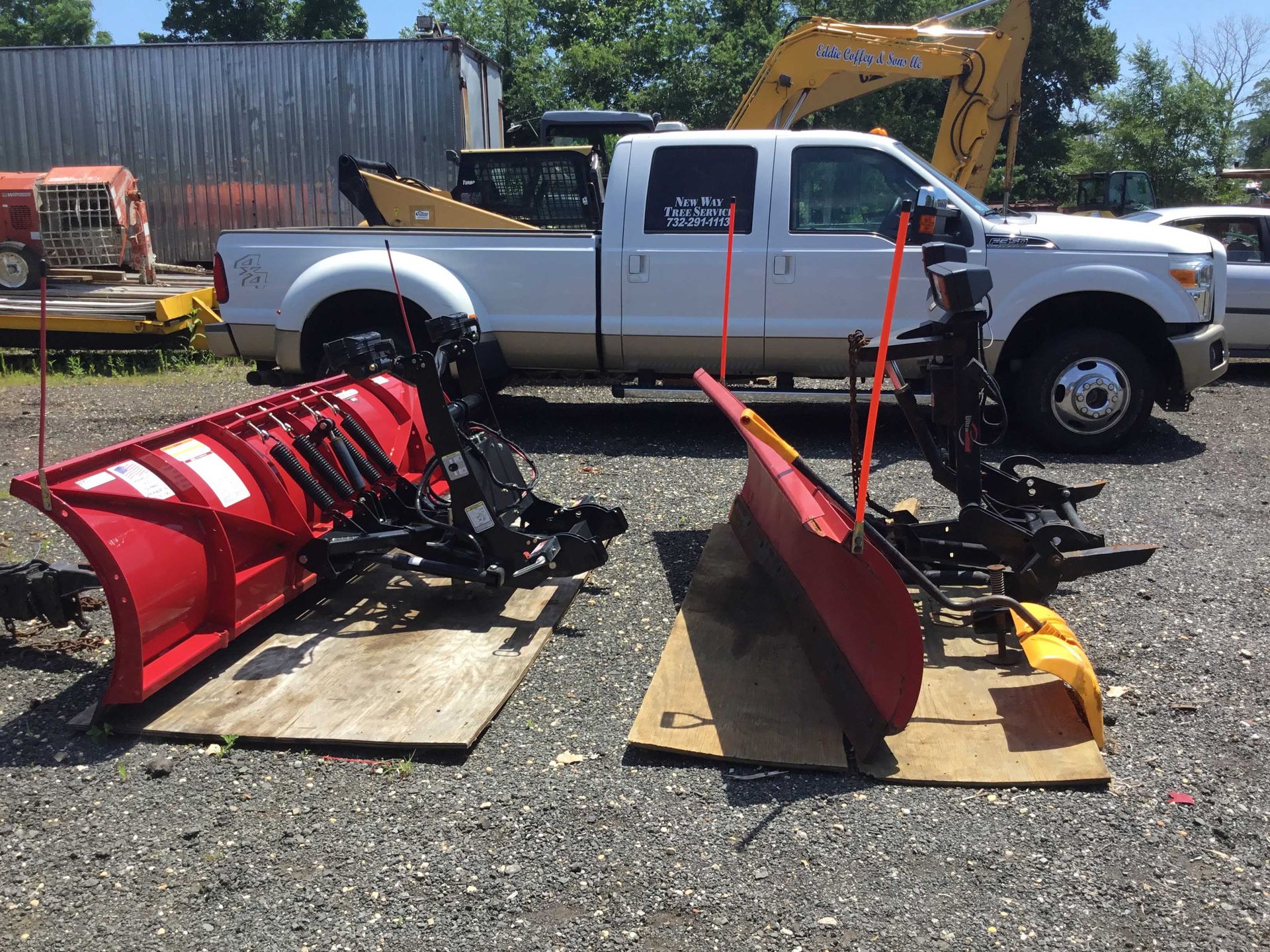 Two red snowplows sit on wooden pallets, beside a white pickup truck on gravel.