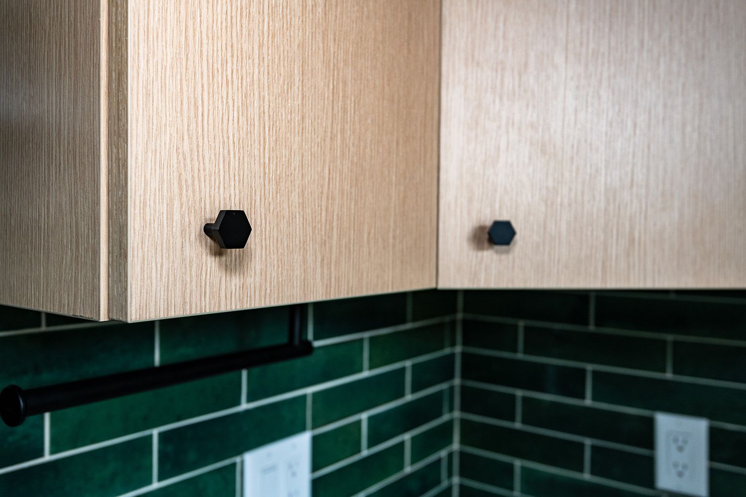 Wooden kitchen cabinets above dark green tile backsplash with black knobs and outlets.