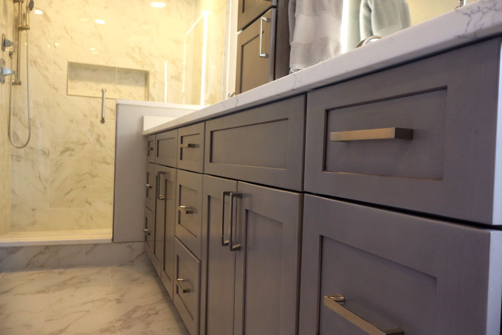 Modern bathroom vanity with gray cabinets, brass handles, and marble shower walls in the background.