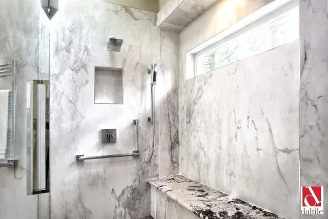 A bathroom with marble walls and a walk in shower with a bench.
