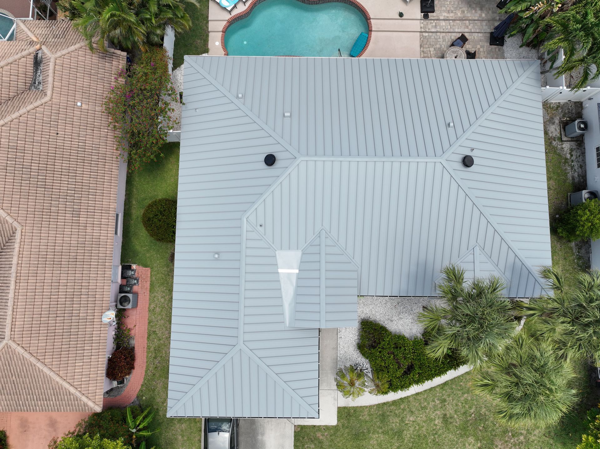 Overhead view of a house with a gray metal roof, a pool, and surrounding green yard and foliage.