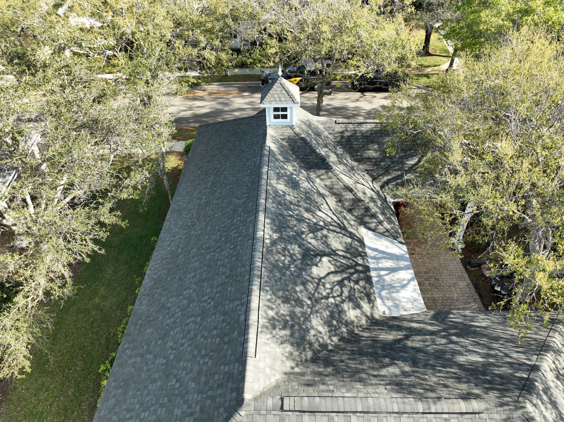 Overhead view of a long, dark gray roof with a white cupola, surrounded by trees on a grassy area.
