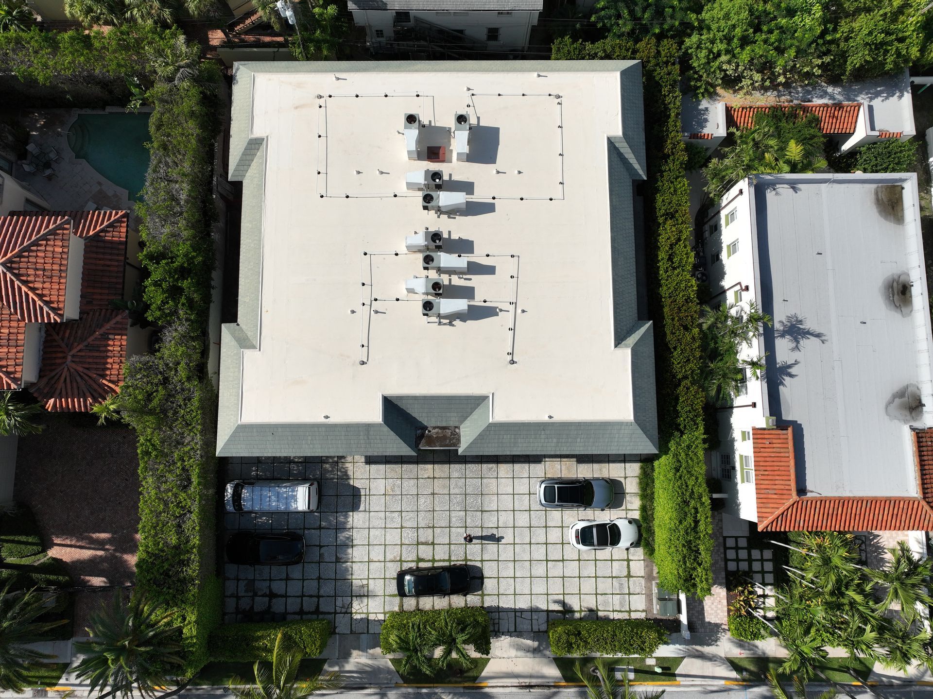 Aerial view of a rectangular building with a flat roof, surrounded by green hedges and adjacent to other buildings. Cars parked in front.