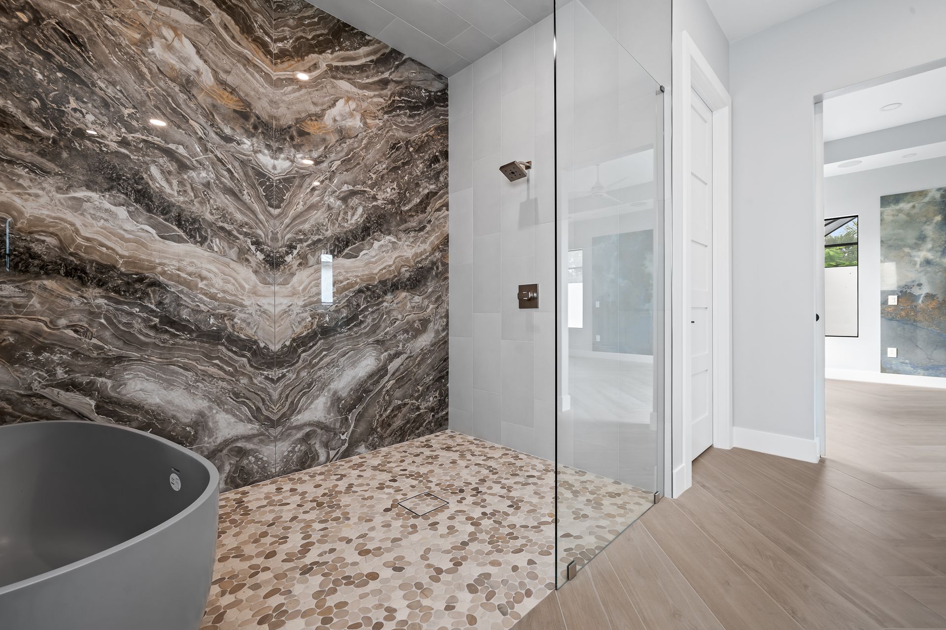 Luxurious bathroom with stone accent wall, glass shower, pebble floor, and gray bathtub.