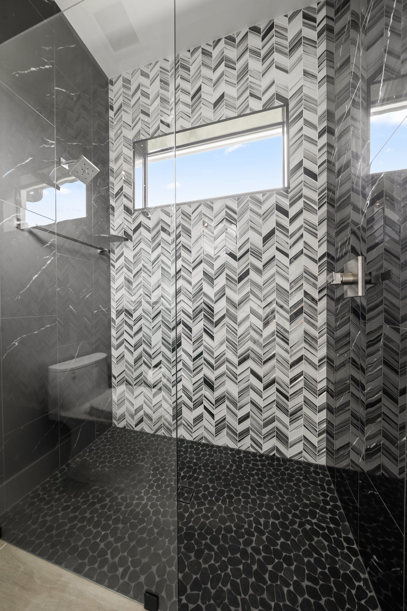 Shower with dark pebble floor, gray and white chevron tile wall, bench, and window.