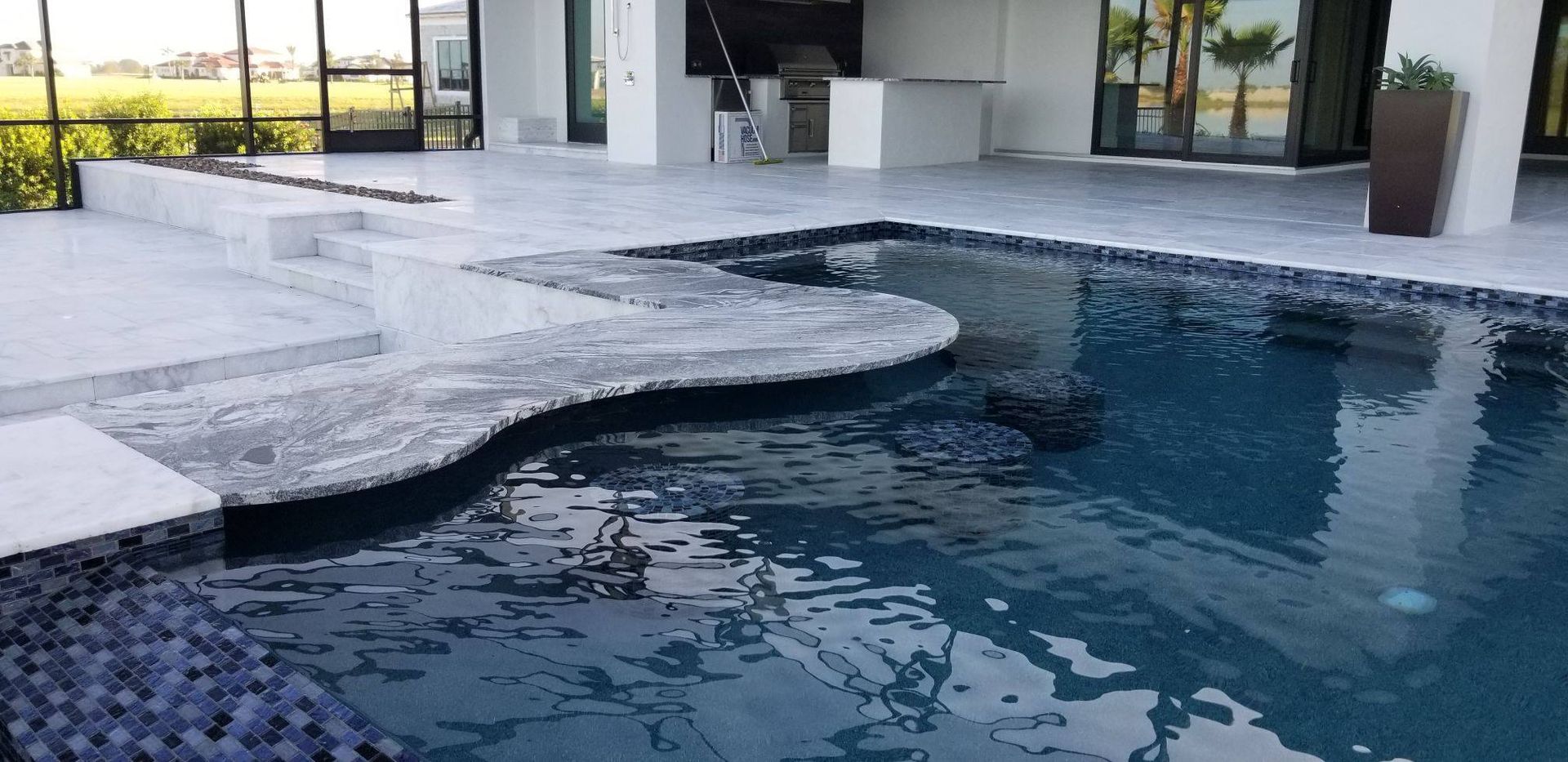 Pool with a tiled edge, surrounded by a white patio with modern architecture in the background.