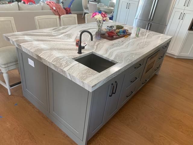 Kitchen island with gray cabinets and white marble countertop, dark sink and faucet, and wooden floor.