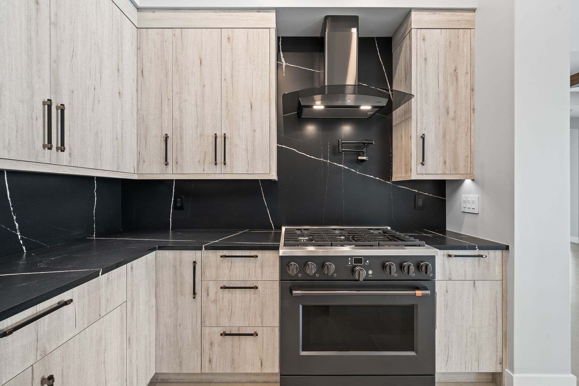 Modern kitchen with light wood cabinets, black countertops, and a stainless steel range and hood.