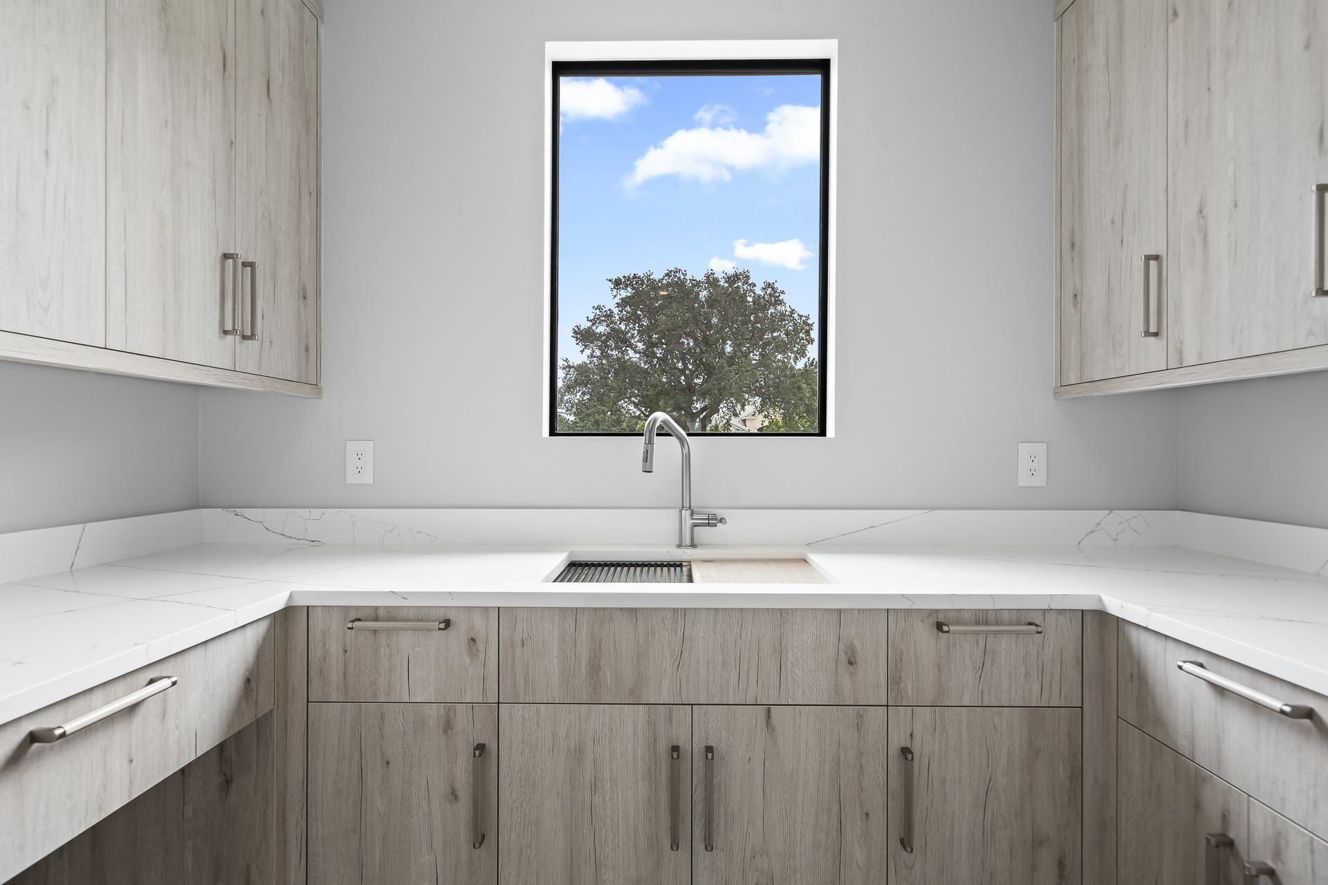 U-shaped laundry room with light wood cabinets, white countertops, and a window with a view of a tree.