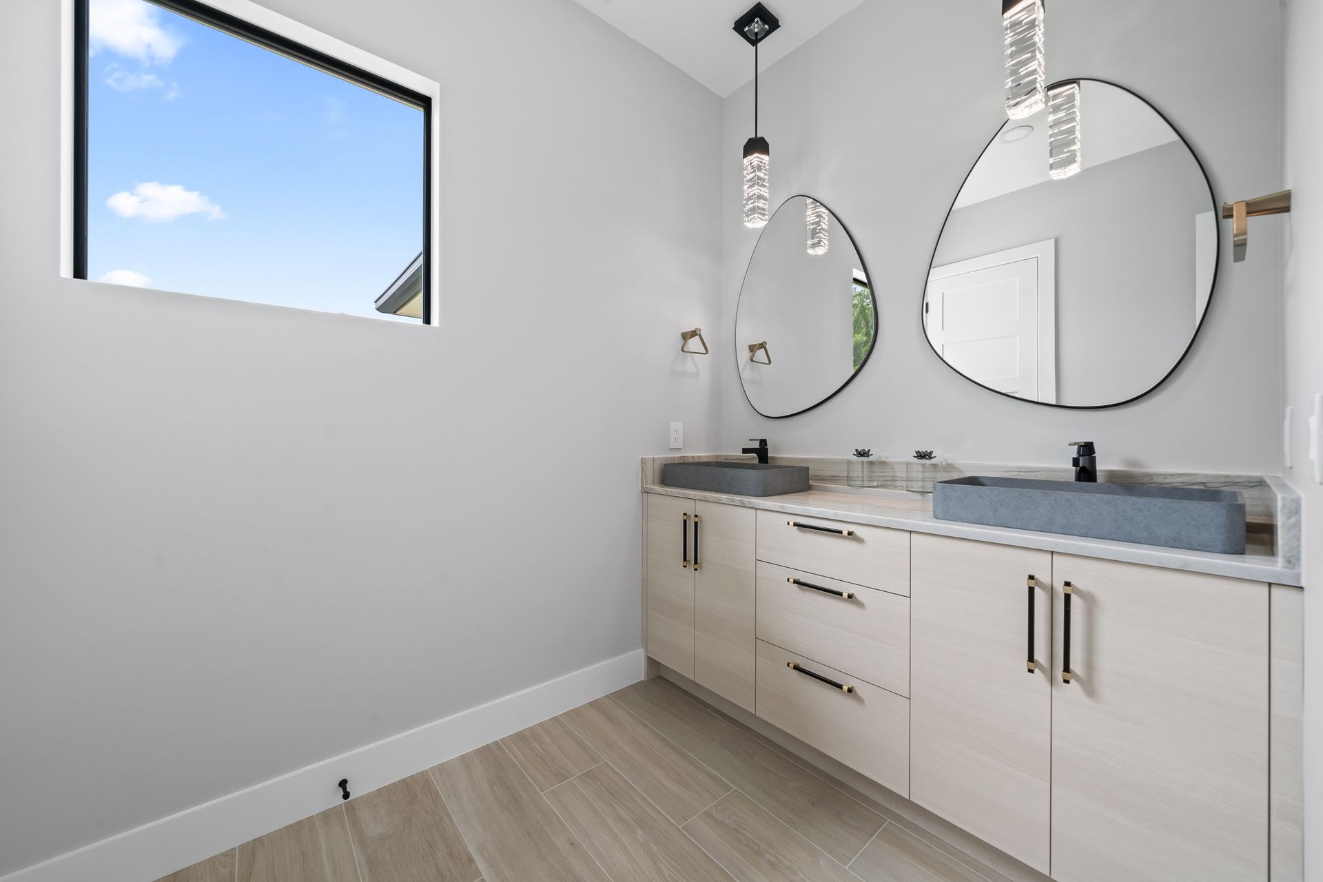 Modern bathroom with two sinks, oval mirrors, and a window with a blue sky view. Light wood cabinets and gray walls.
