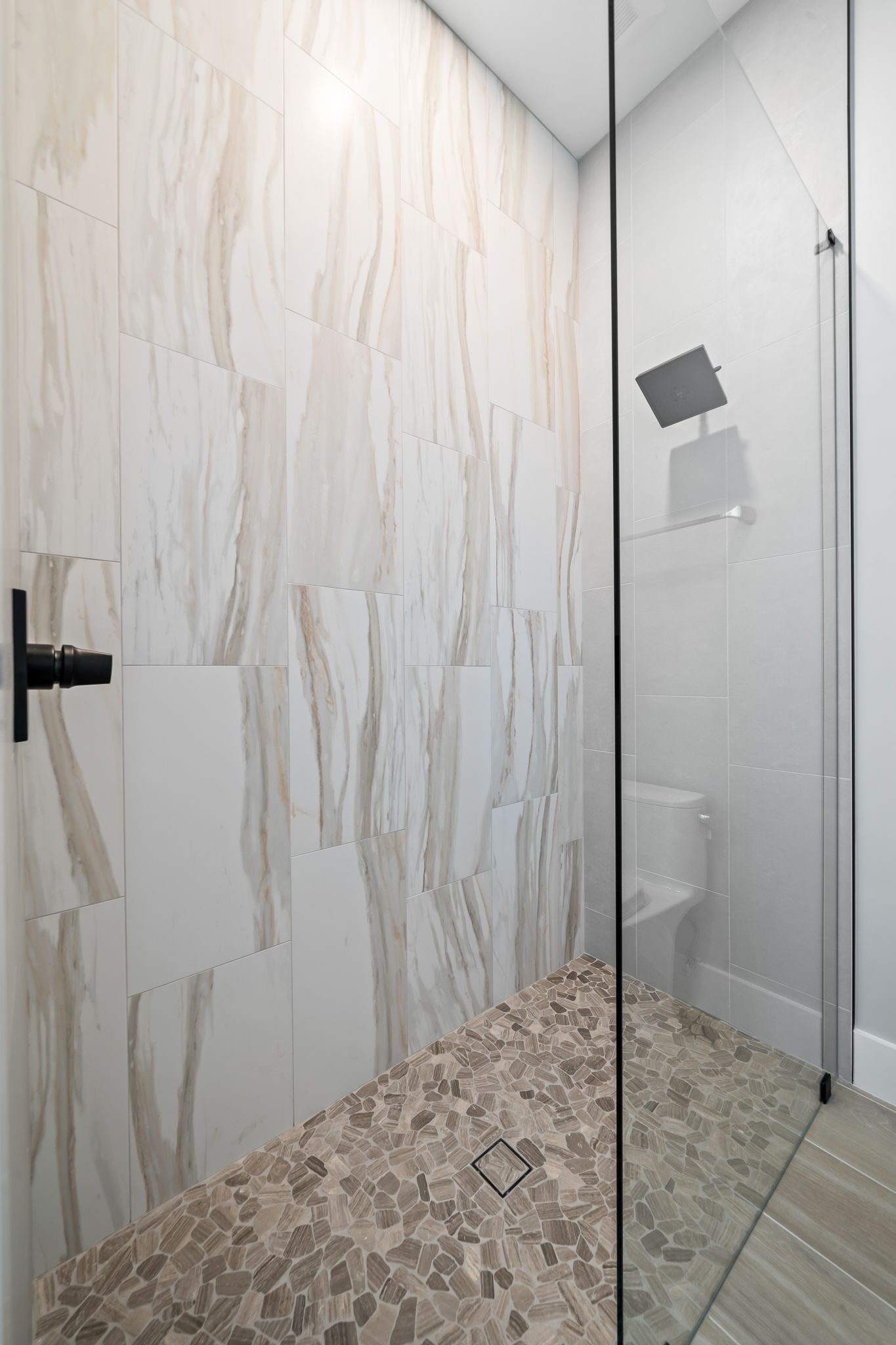 Shower stall with pebble floor and stone-patterned wall tiles. Clear glass door and black fixtures.
