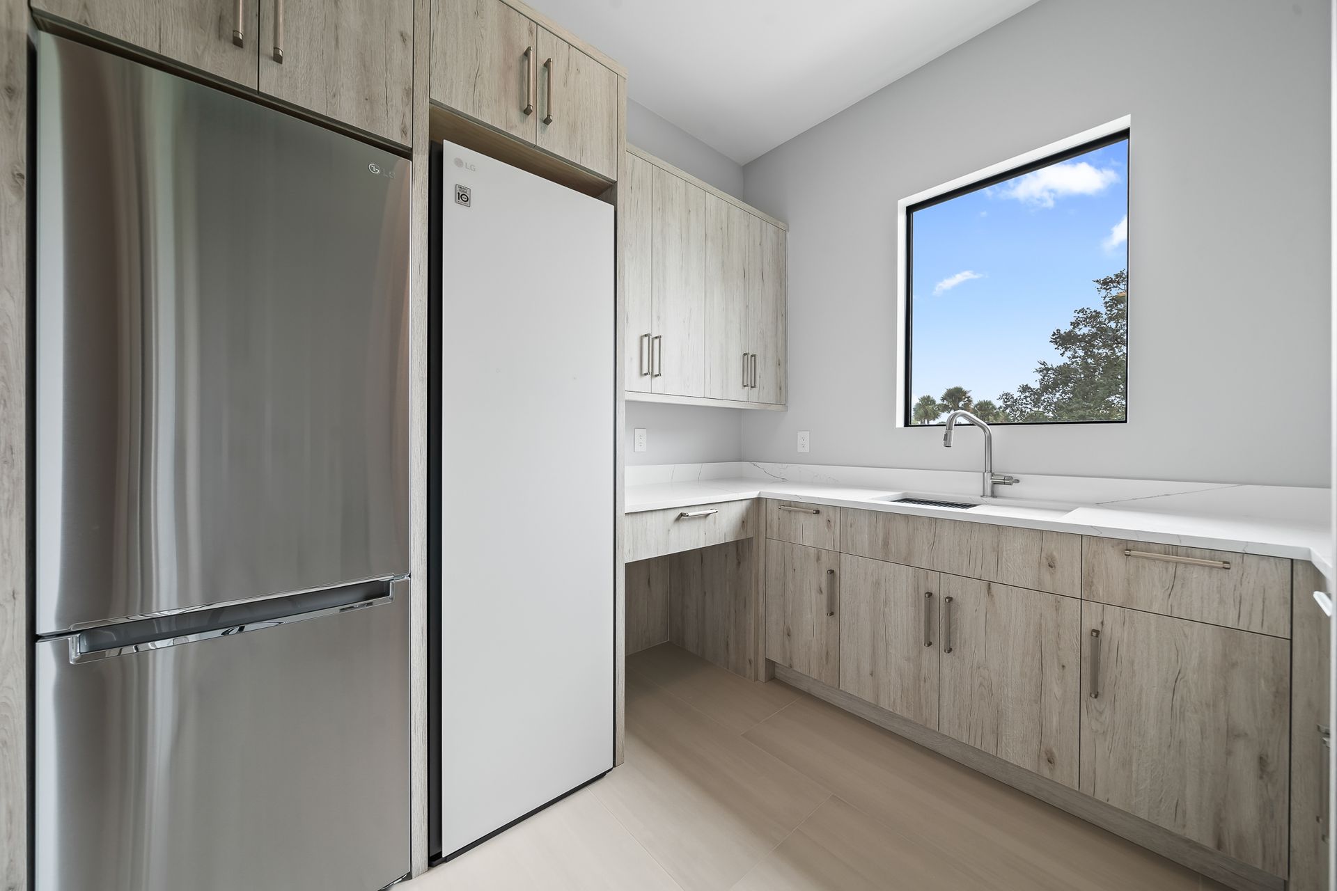 Modern kitchen with light wood cabinets, stainless steel refrigerator, white countertops, and window overlooking trees.