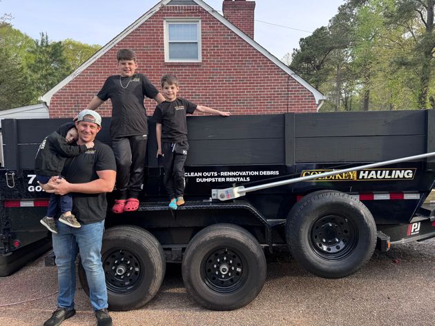 Four people posing on a black dump trailer in front of a red brick house