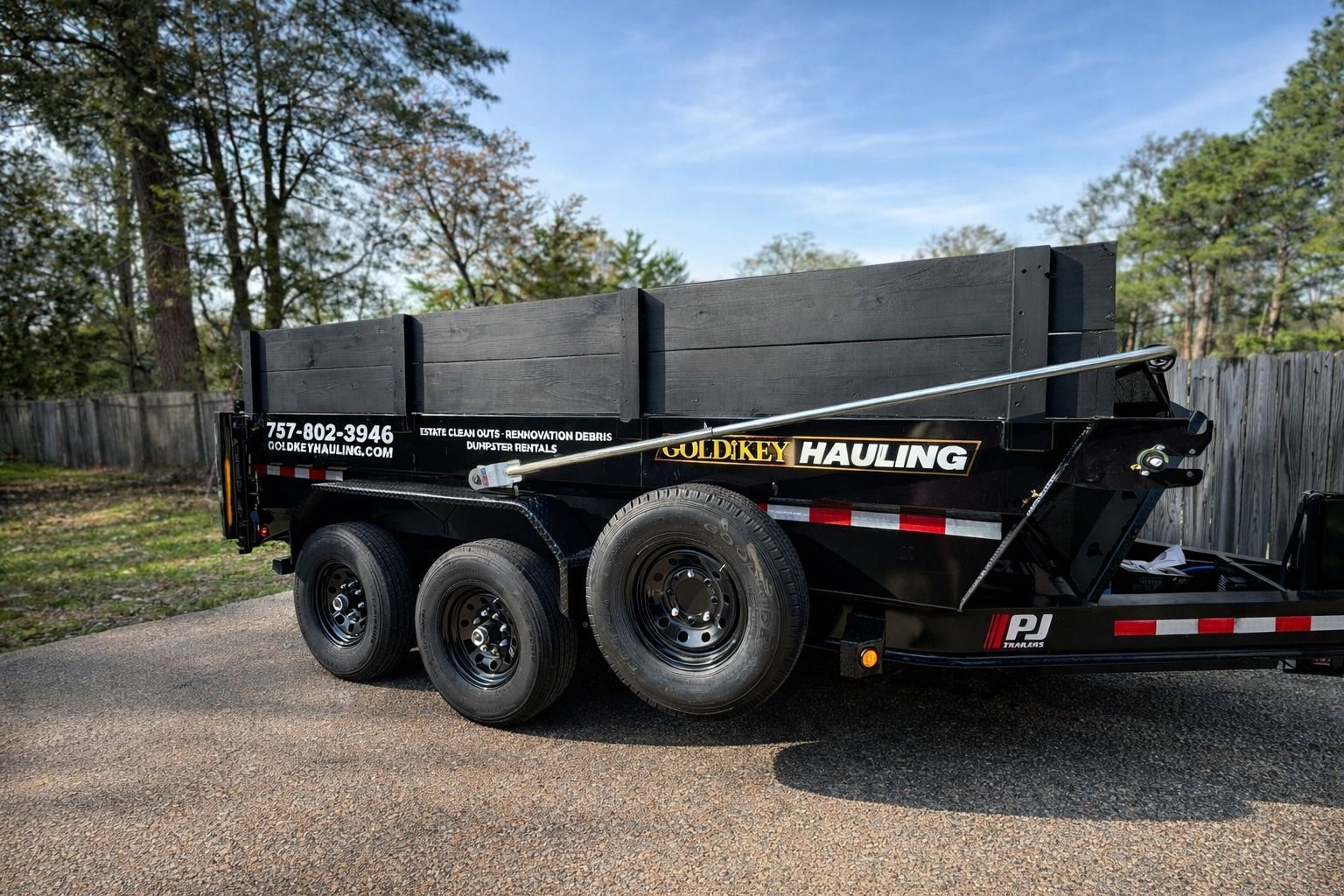 Black dump trailer with three axles parked on gravel near trees and a blue sky.