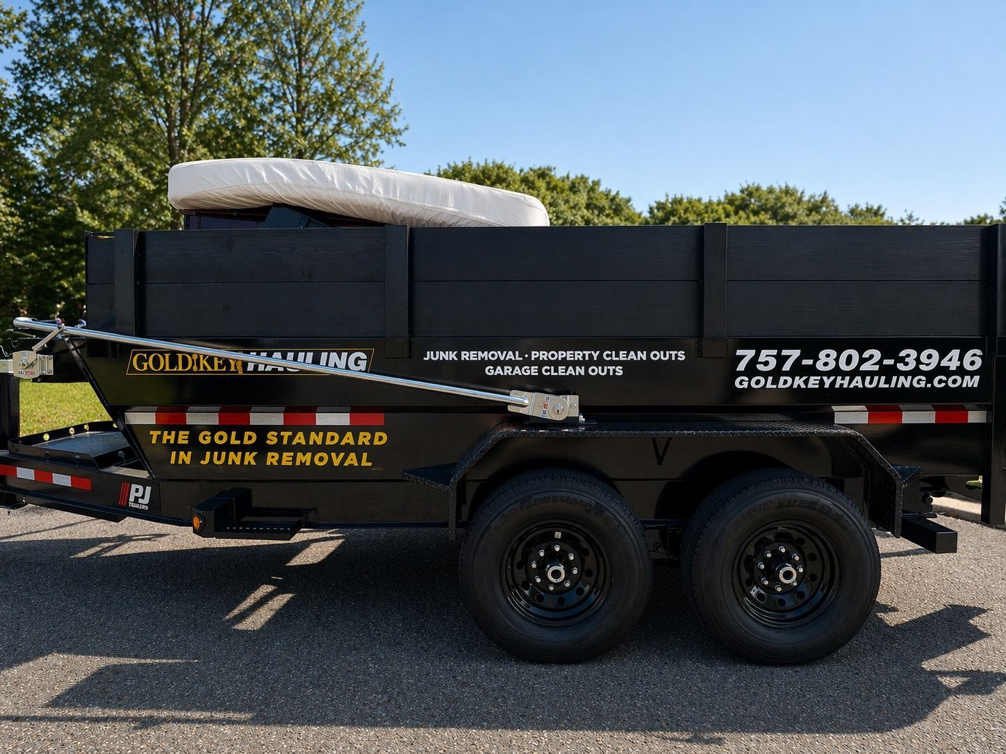 Black dump trailer with company decals and phone number, parked on gravel outdoors.
