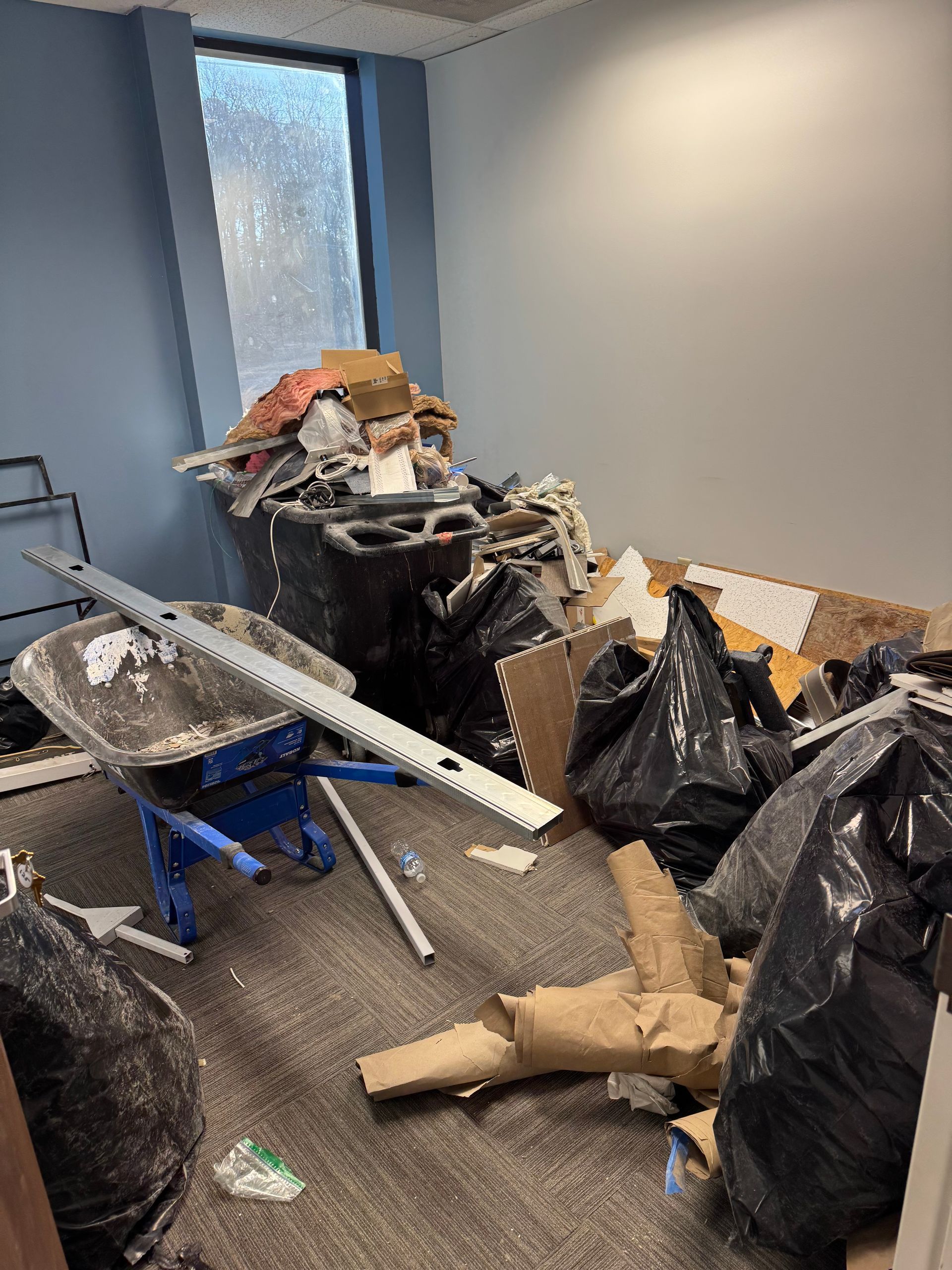 Office hallway cluttered with trash bags, cardboard, and broken furniture near a window. - before