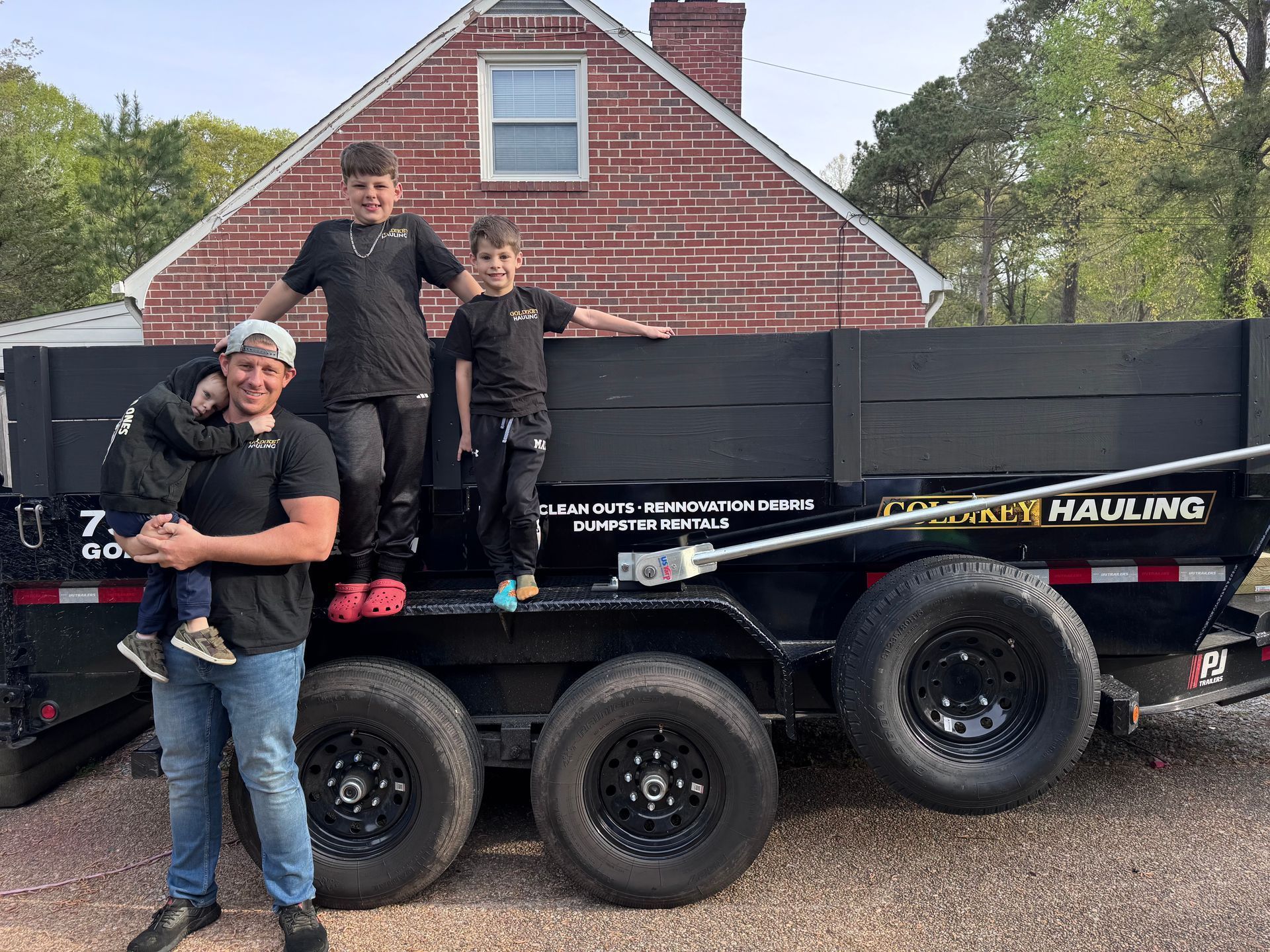 Three people posing on a black dump trailer with a red brick house in the background