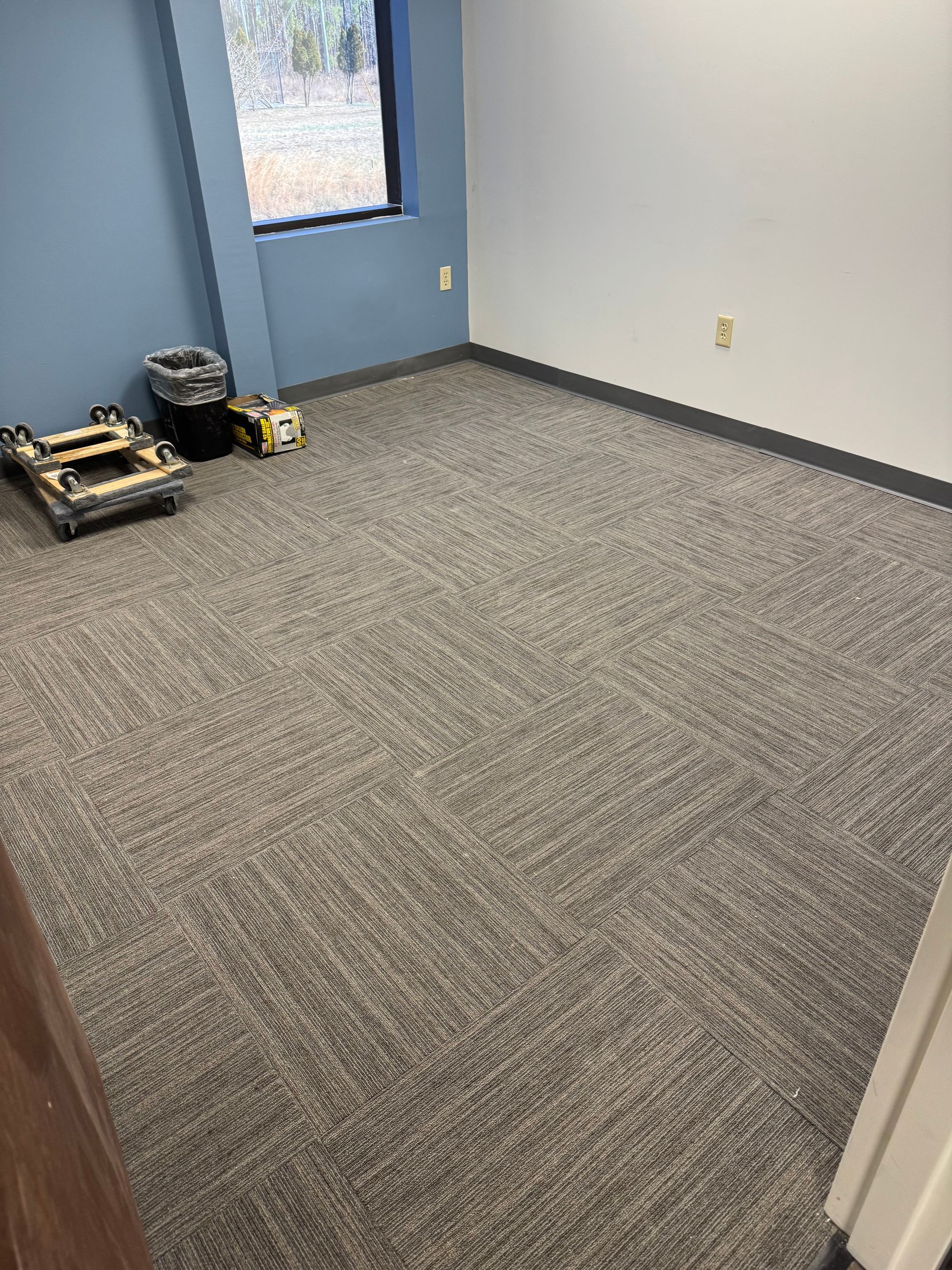 Empty office with gray carpet, light blue wall, window, and a few items stacked in the corner. - after