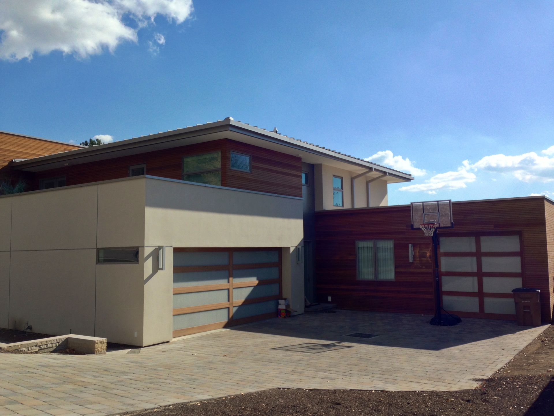Modern suburban house with beige and wood exterior, garage doors, and a paved driveway under a blue sky