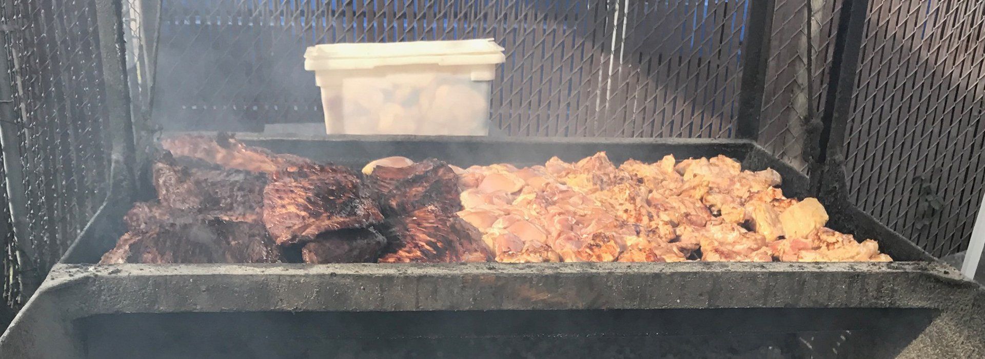 Smoke rising over a grill with charred meat on the left and golden chicken pieces on the right, with a tub in back