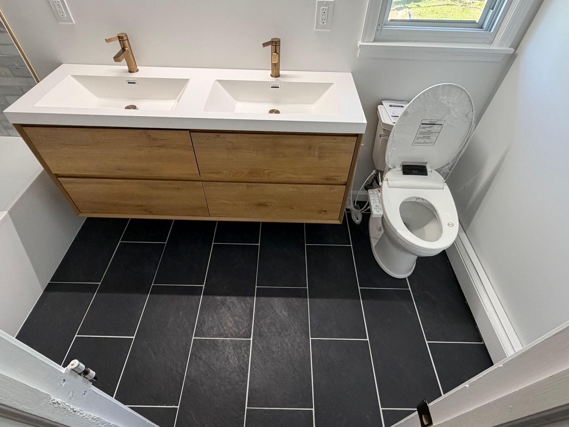 Modern bathroom with double sink vanity, black tile floor, and toilet under a small window.