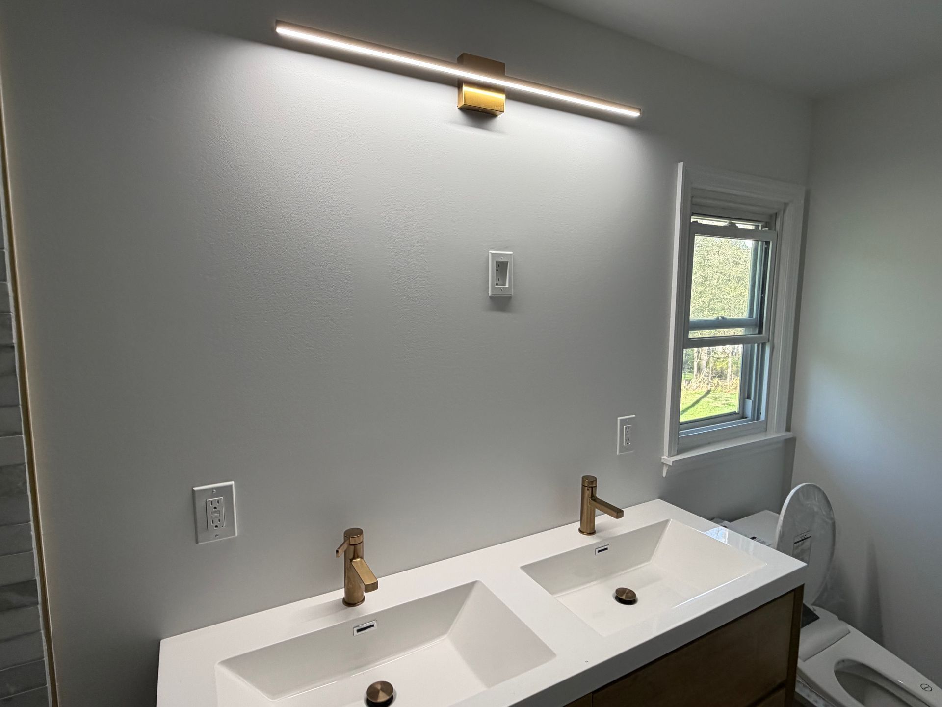 Modern bathroom vanity with two sinks, brass faucets, wall light, and a window