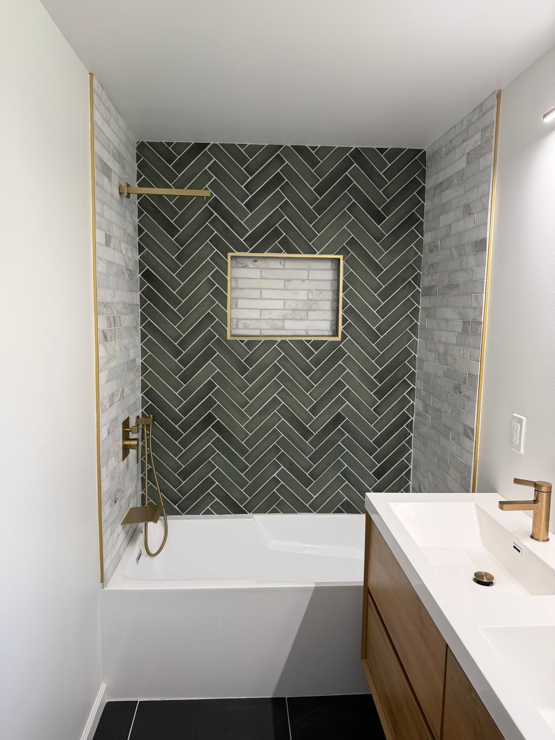 Modern bathroom with green herringbone tile, white tub, and wood vanity with gold fixtures