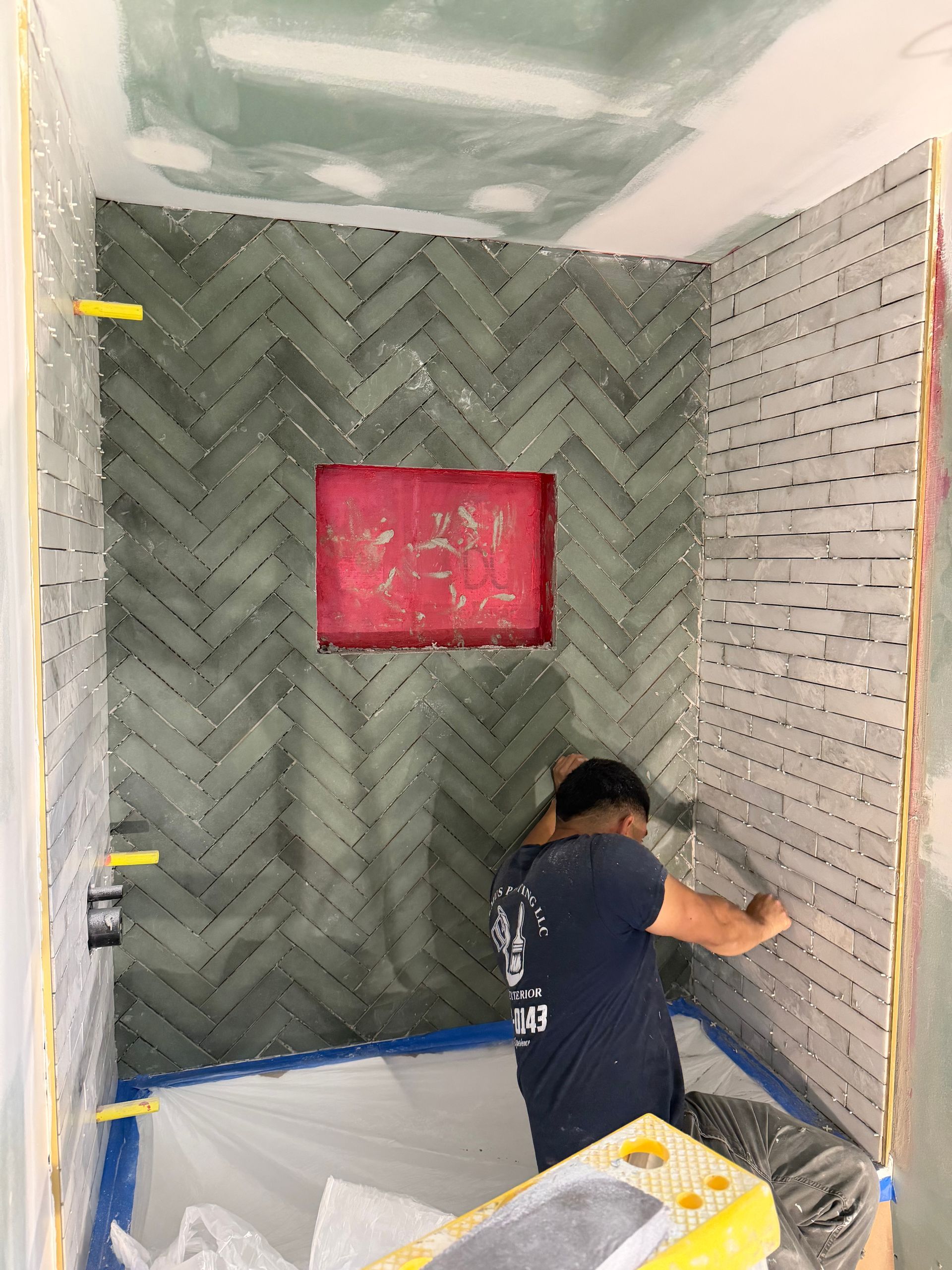 Worker tiling a shower alcove with gray herringbone tile and a pink niche shelf