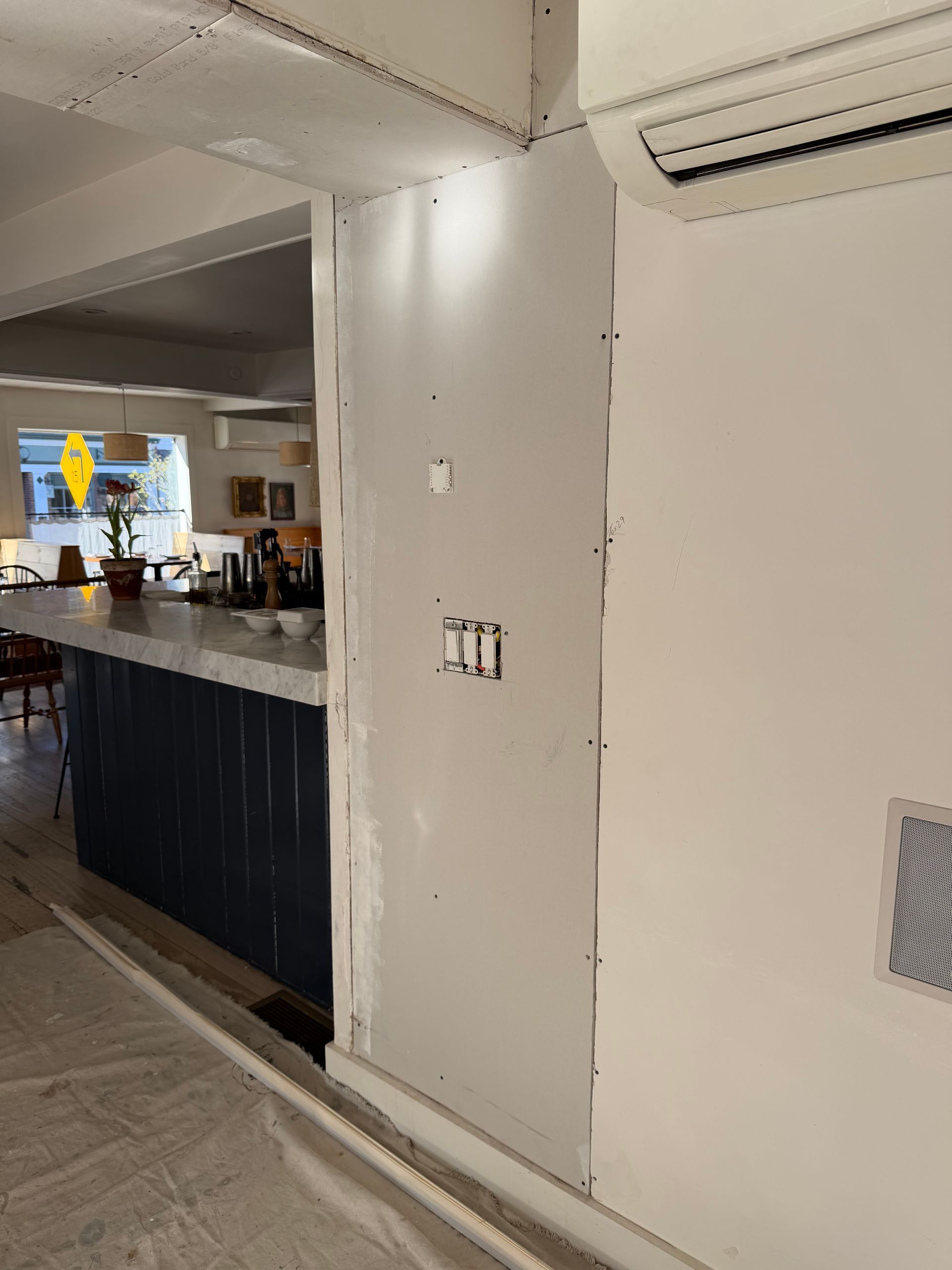 A wall under renovation with exposed drywall, a light switch opening, and an adjacent kitchen view.
